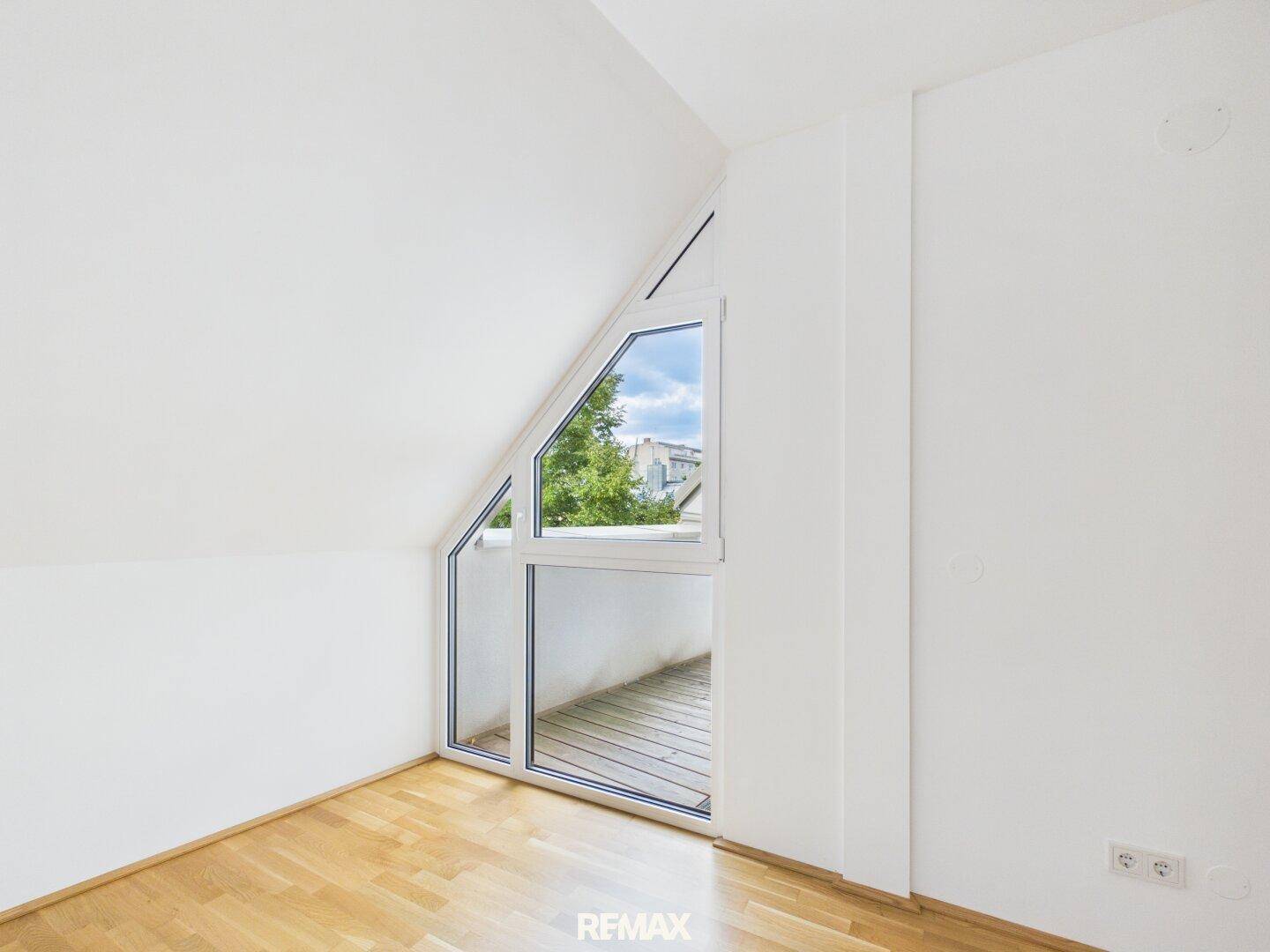 Dachzimmer mit schräger Decke und Fenster, das Zugang zu einem kleinen Balkon bietet.