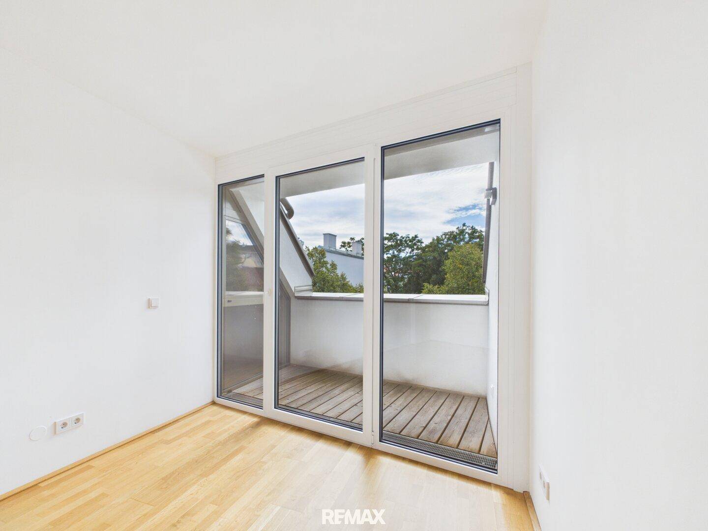 Leeres Zimmer mit Parkettboden und großen Fenstern, die zu einem kleinen Balkon führen.