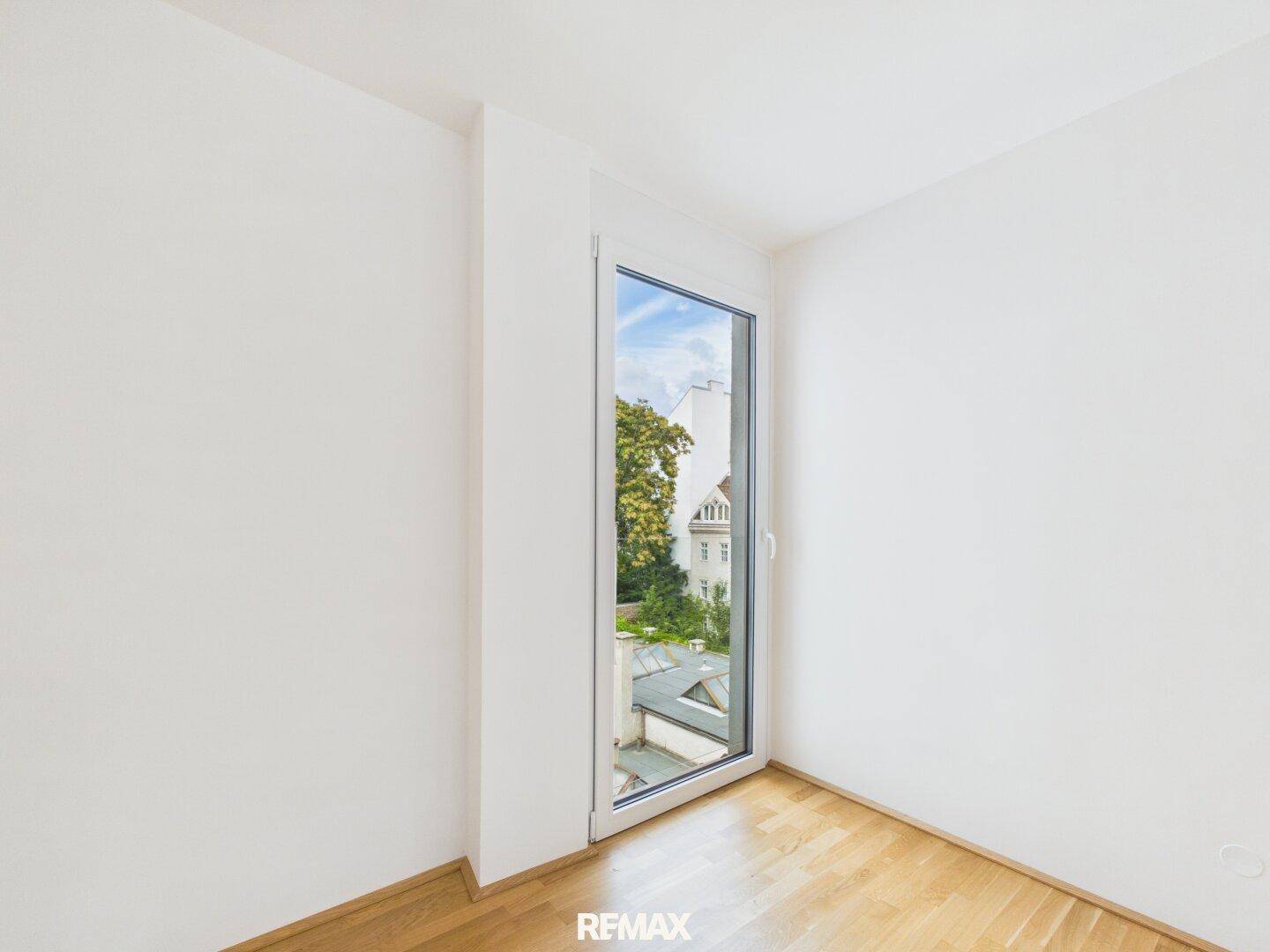 Helles Zimmer mit Parkettboden und einem hohen Fenster, das einen Blick auf die Stadt ermöglicht.