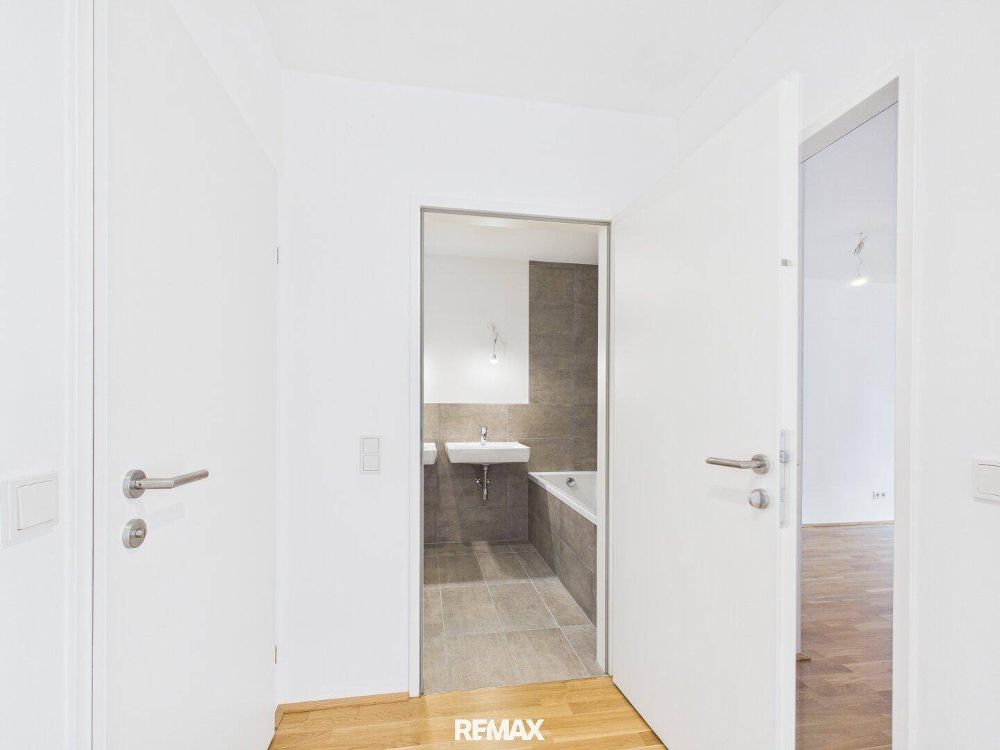 Modernes Badezimmer mit Badewanne, Waschbecken und hellen Fliesen, sichtbar durch eine offene Tür.