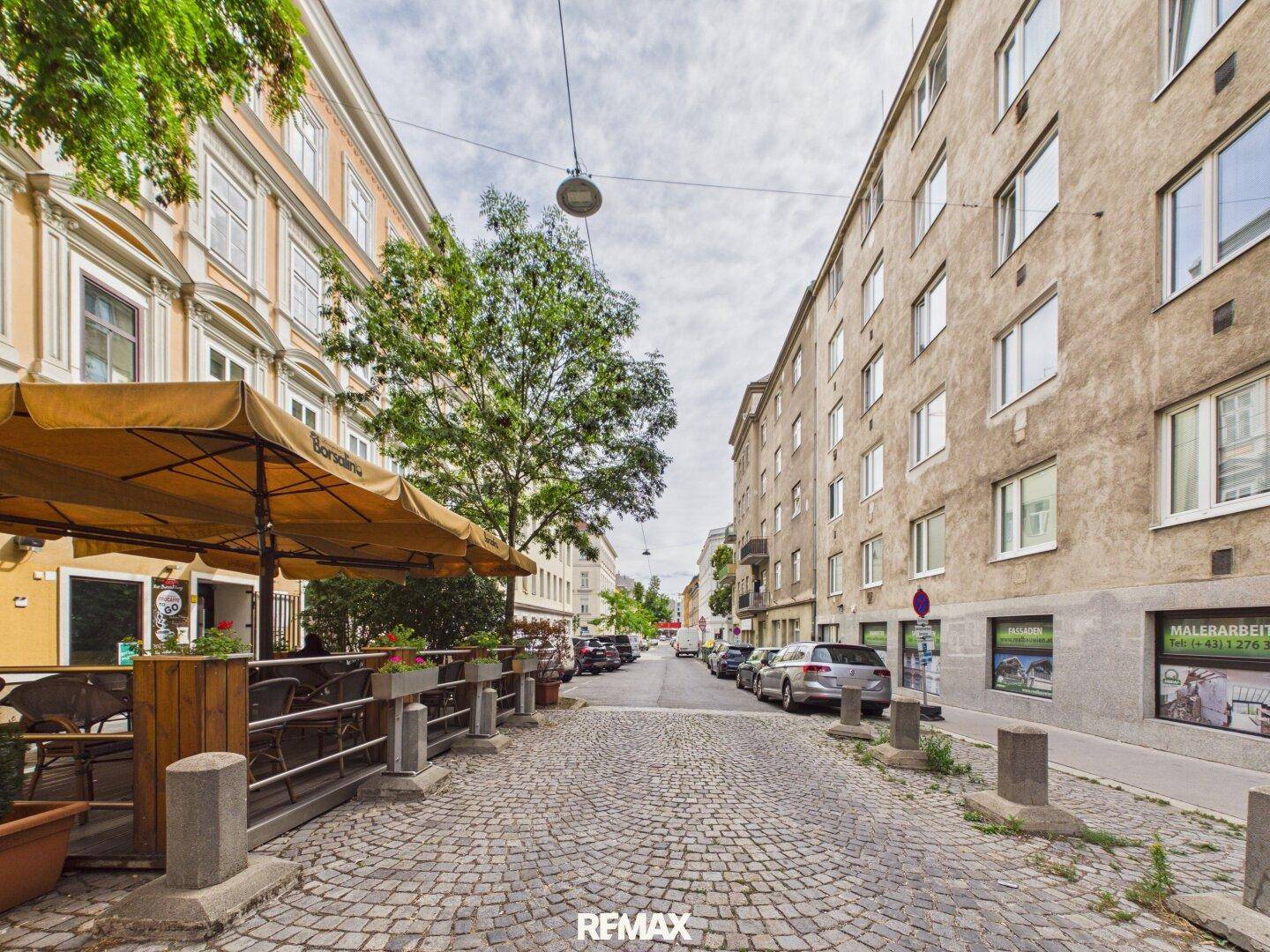 Kopfsteingepflasterte Straße mit Bäumen, Cafés und geparkten Autos in der Stadt.
