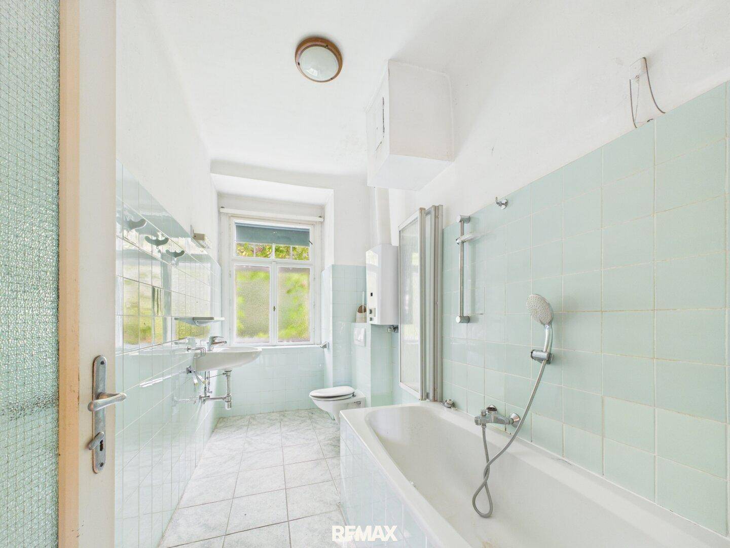 Badezimmer mit hellblauen Fliesen, Badewanne und Fenster, renovierungsbedürftig.