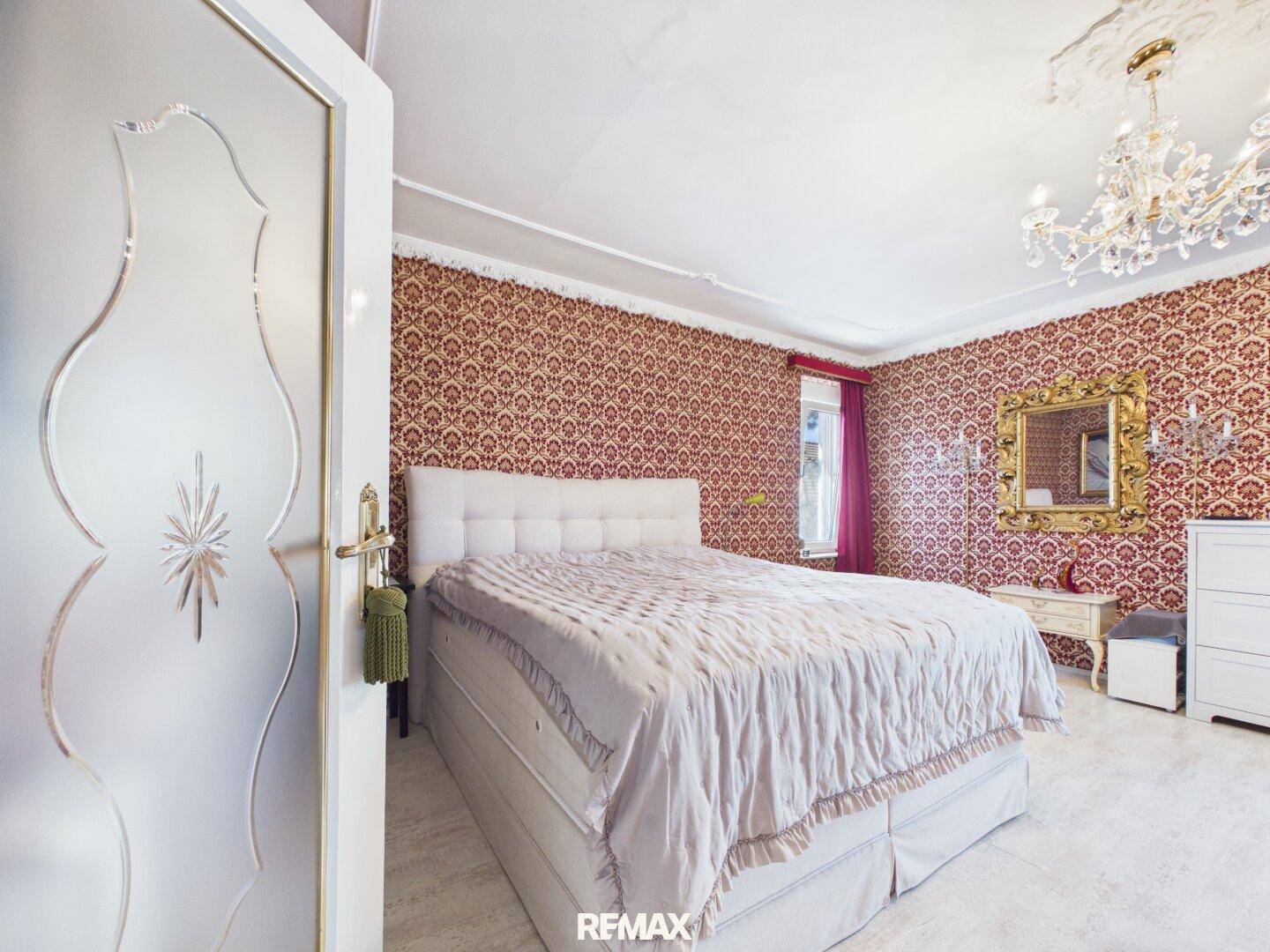 Elegantes Schlafzimmer mit großem Bett, gemusterter Tapete, Kronleuchter und Goldspiegel.
