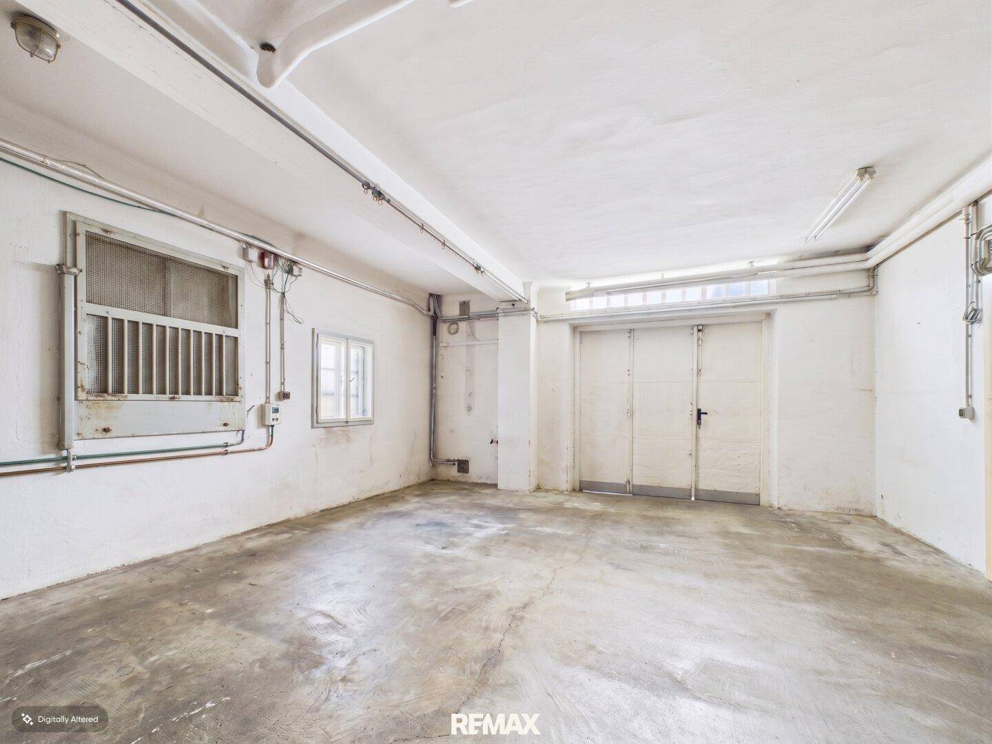 Geräumiger leerer Raum mit Betonboden, zwei Fenstern und großen Doppeltüren, ideal als Garage oder Lager.