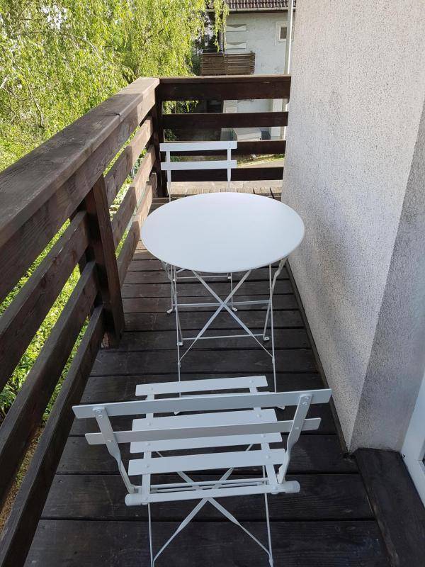 Kleiner Balkon mit Holzboden und weißen Gartenmöbeln, umgeben von viel Grün.