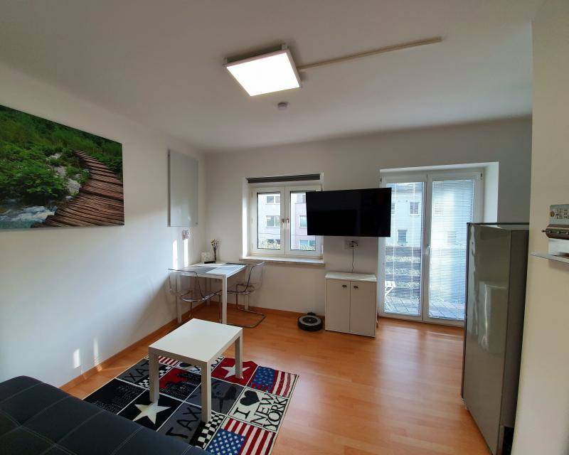 Geräumiges Wohnzimmer mit Holzboden, Fernseher und einem Kühlschrank im Hintergrund.