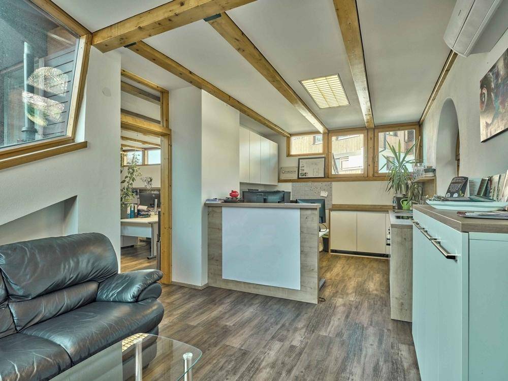 Einladender Empfangsbereich mit Ledersofa, Holzbalken und großen Fenstern, die viel Tageslicht hereinlassen.