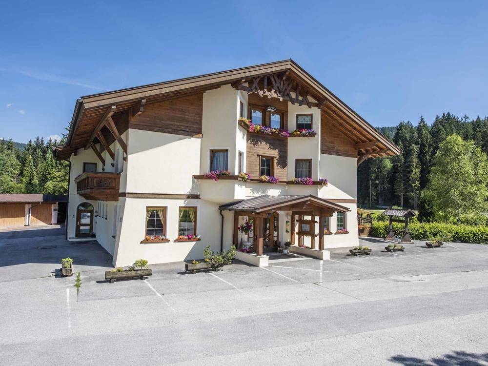 Traditionelles Gebäude im alpinen Stil mit Holzbalkonen und Blumenkästen, umgeben von Bäumen und Bergen.