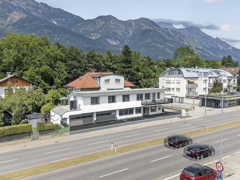 Panoramablick auf das moderne Gebäude an der Hauptstraße, umgeben von üppiger Natur und einer beeindruckenden Bergkette.