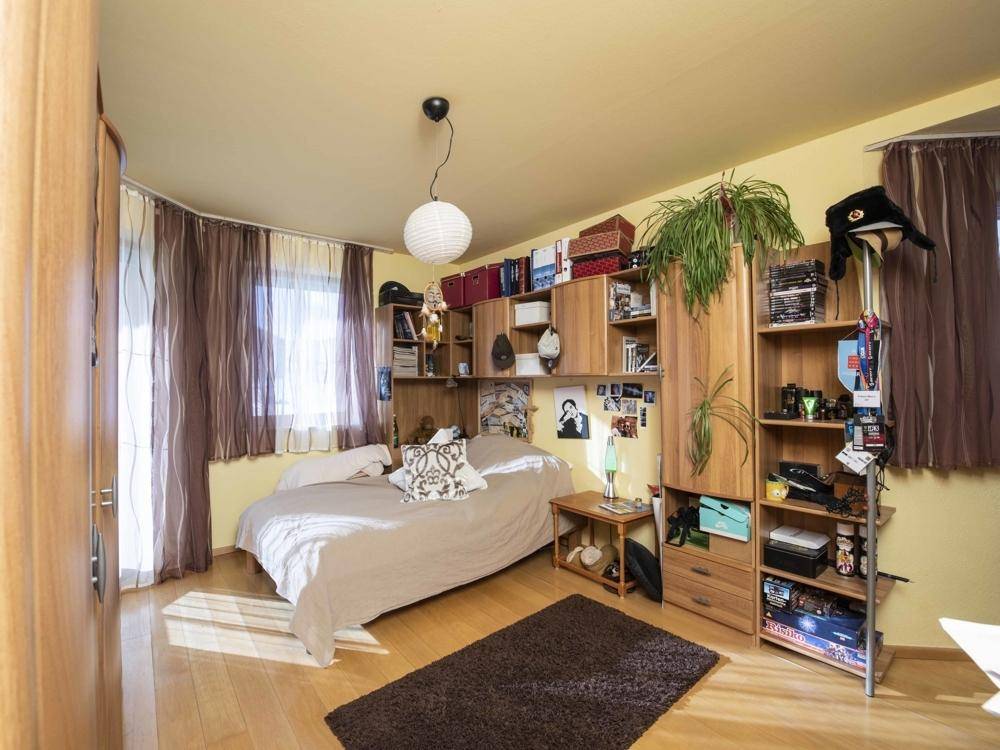 Gemütliches Schlafzimmer mit Holzboden, Bücherregalen und einem Fenster mit braunen Vorhängen.