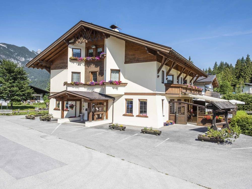 Traditionelles Gebäude im alpinen Stil mit Holzbalkonen und Blumenkästen, umgeben von Bäumen und Bergen.