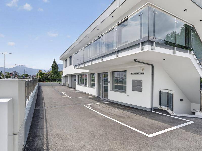 Ansicht des modernen Gebäudes mit Eingangsbereich, markierten Parkplätzen und einer eleganten Glasbrüstung.
