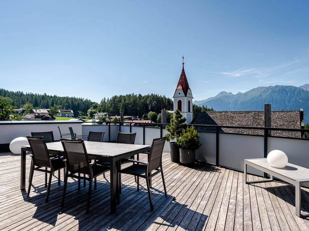 Sonniger Terrassenbereich mit Esstisch und Lounge-Möbeln, bietet einen weiten Blick auf die Berge und das Dorf.