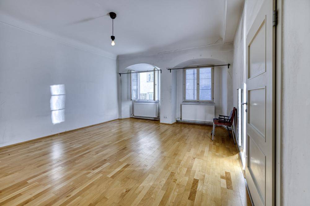 Geräumiges Wohnzimmer mit Holzboden, zwei Fenstern und weiß gestrichenen Wänden.