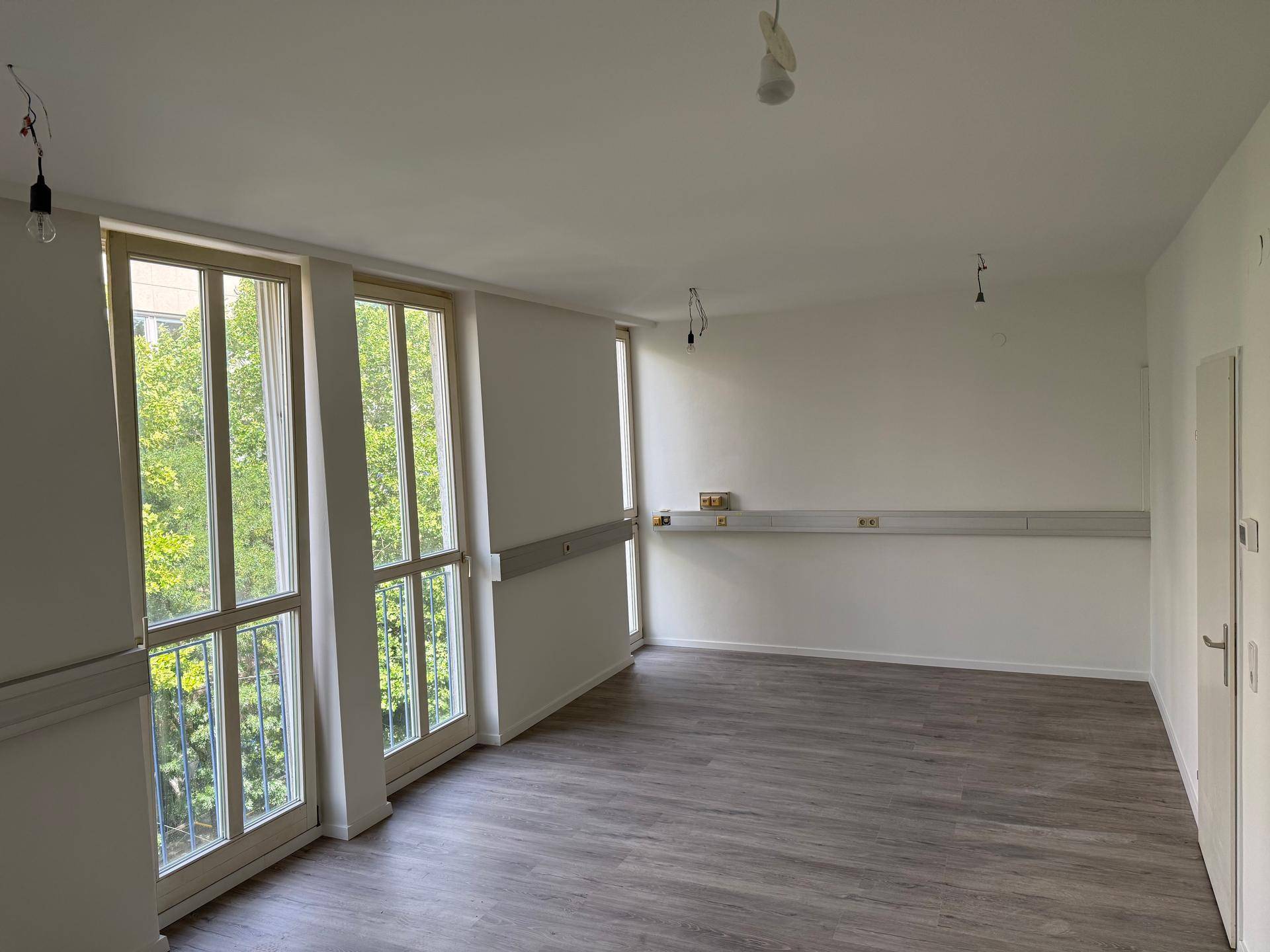 Heller Raum mit Laminatboden und großen Fenstern, die einen grünen Ausblick bieten.