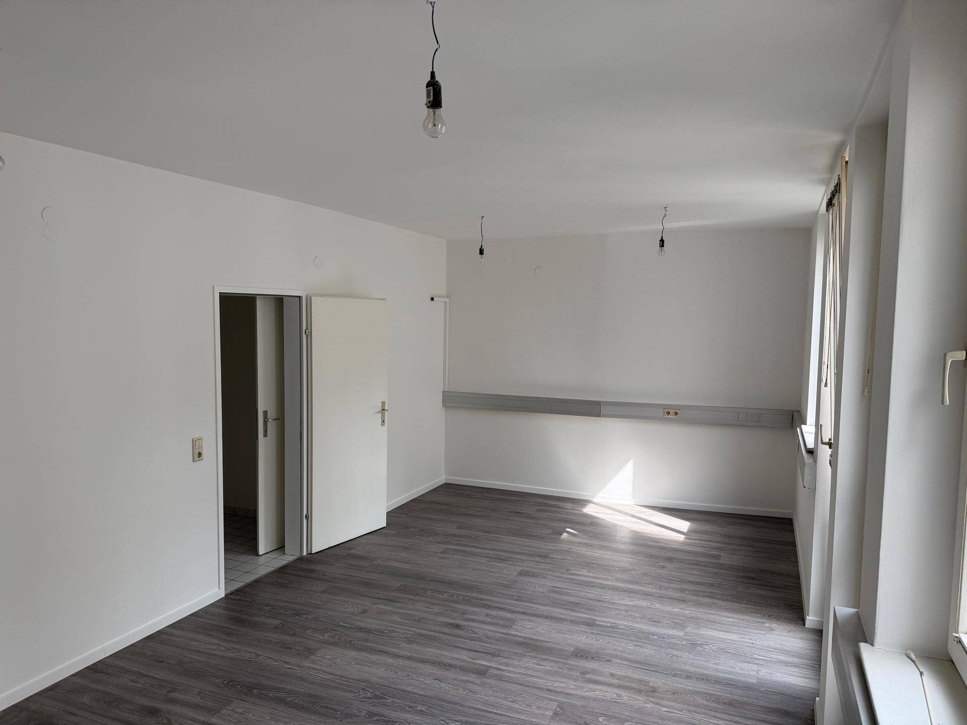 Heller Raum mit Laminatboden und weißen Wänden, durchflutet von natürlichem Licht durch die Fenster.