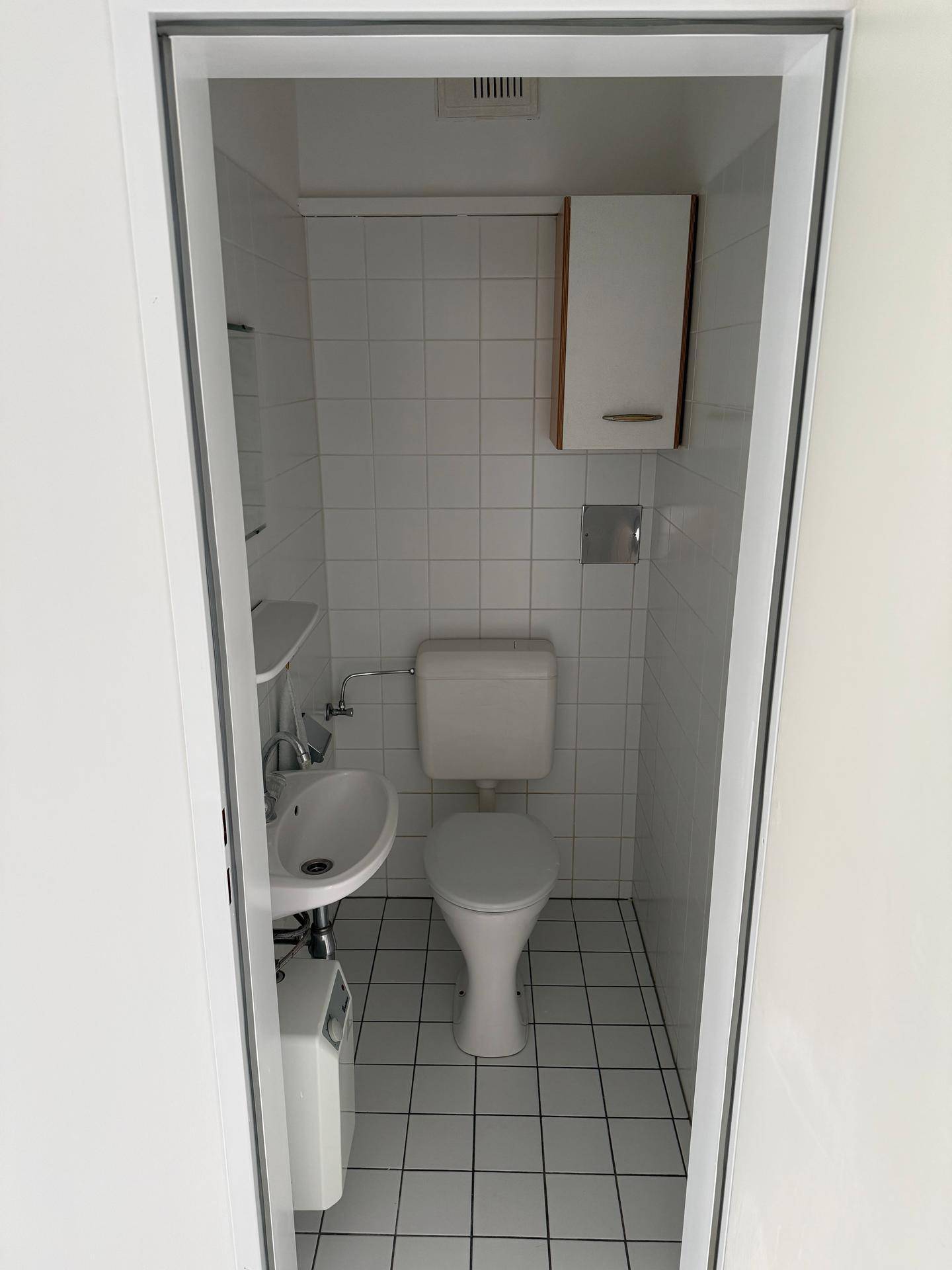 Kompakte Gästetoilette mit weißen Fliesen, kleinem Waschbecken und einem Hängeschrank.