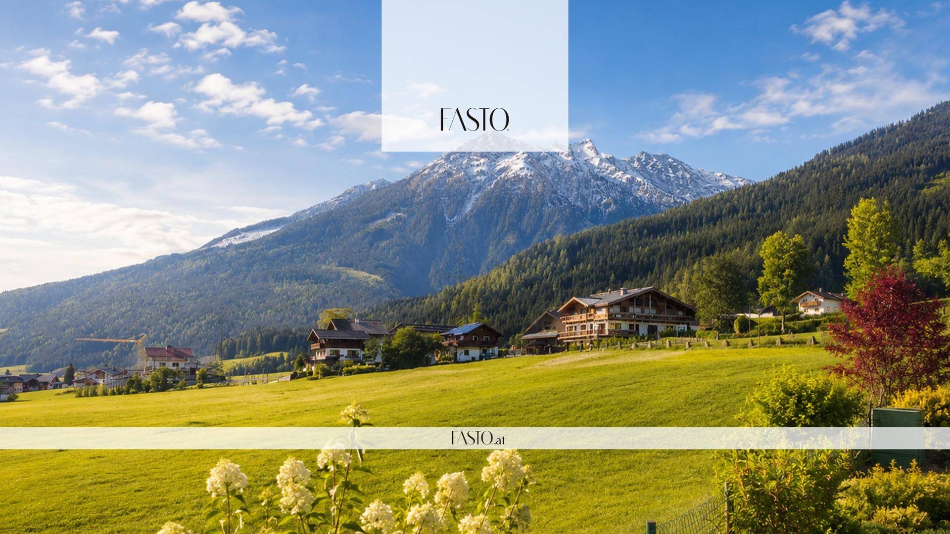 Weitläufige Landschaft mit grünen Wiesen, traditionellen Häusern und schneebedeckten Bergen unter blauem Himmel.