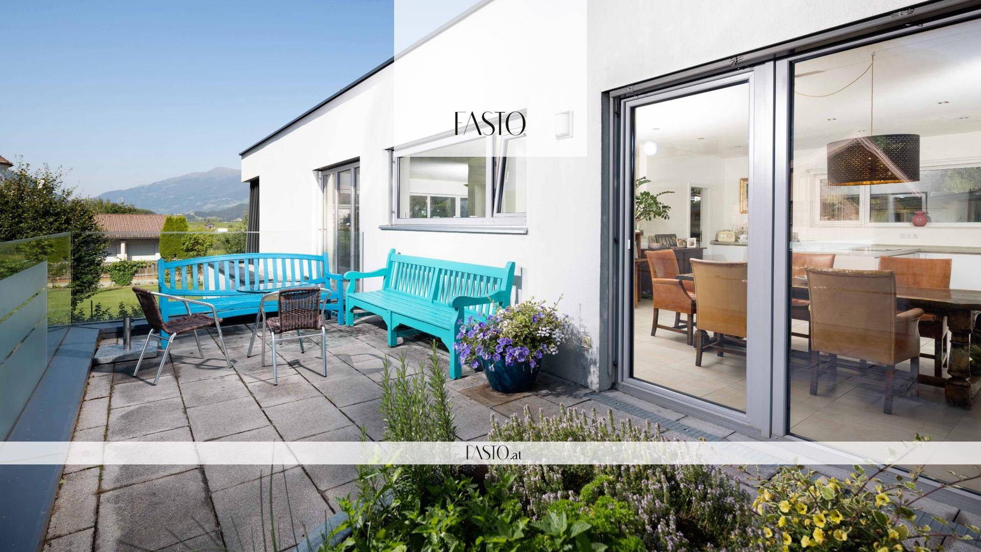 Gepflasterte Terrasse mit blauen Gartenbänken und Blick in den hellen Essbereich.