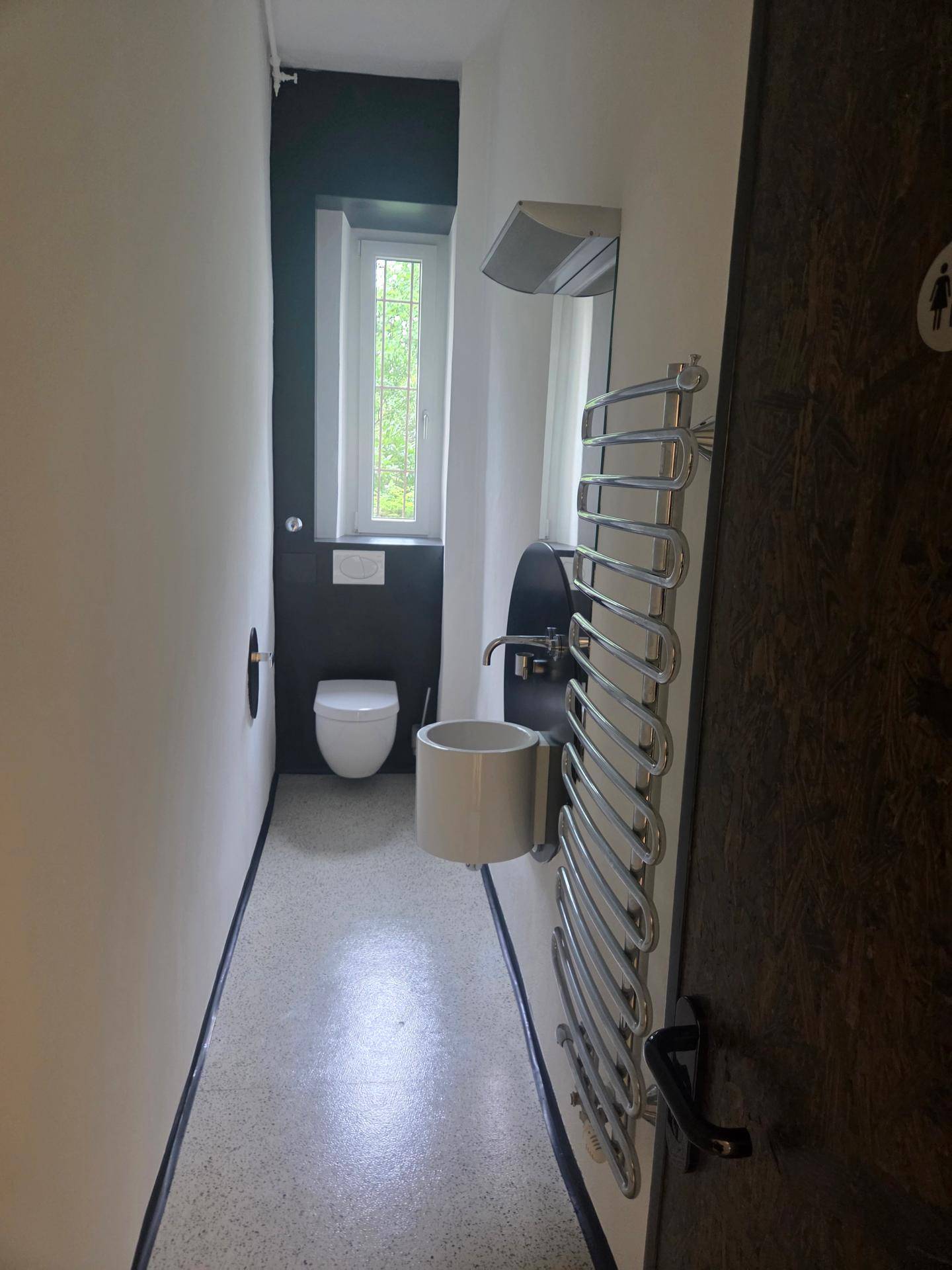 Schmaler Toilettenraum mit modernem Wand-WC, kleinem Handwaschbecken, Handtuchheizkörper und einem Fenster für natürliches Licht.