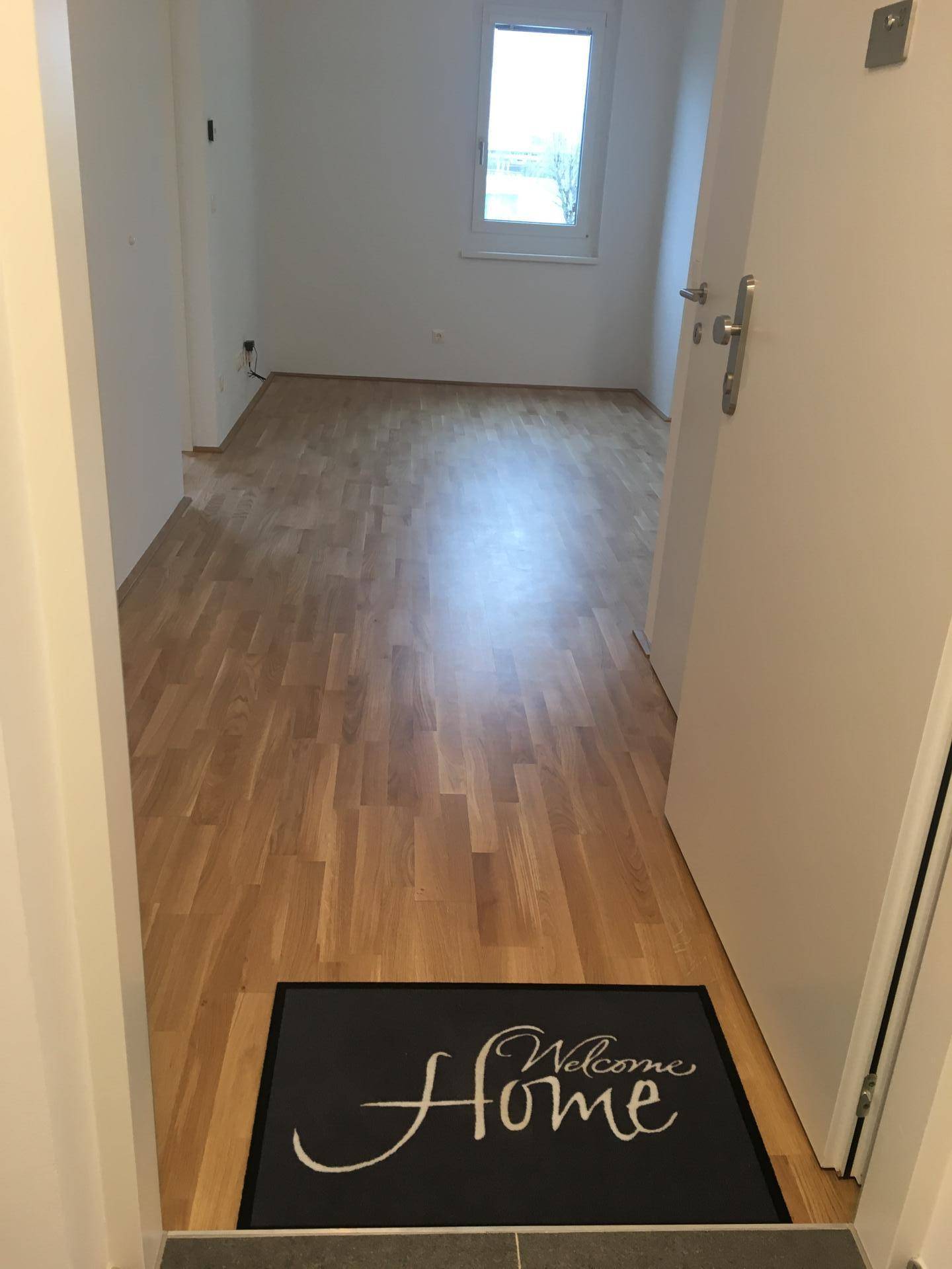 Heller Eingangsbereich mit Laminatboden und einer "Welcome Home" Fußmatte.