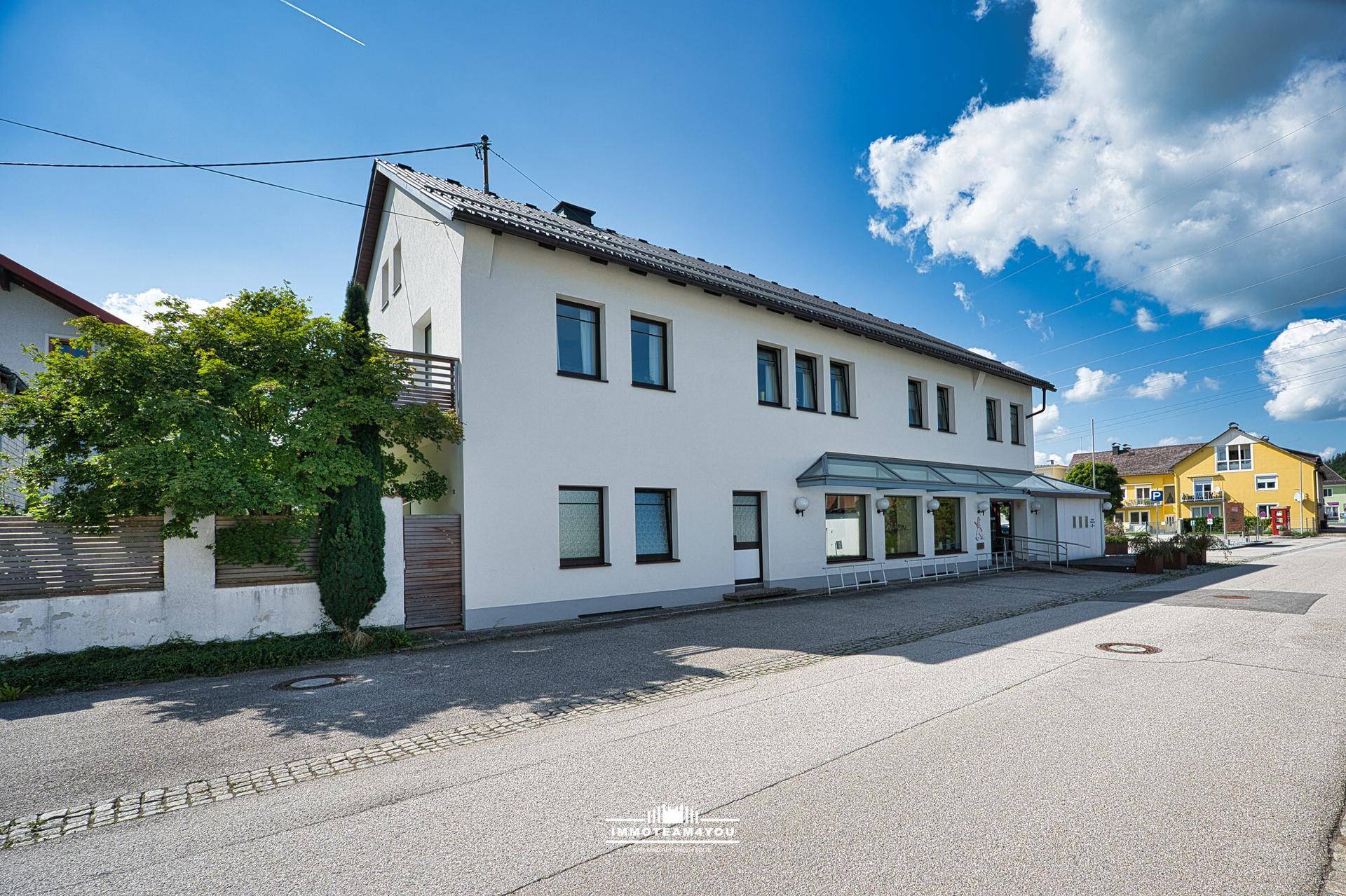 Seitenansicht eines weißen Gebäudes mit Balkon und Zugang zur Straße an einem sonnigen Tag.