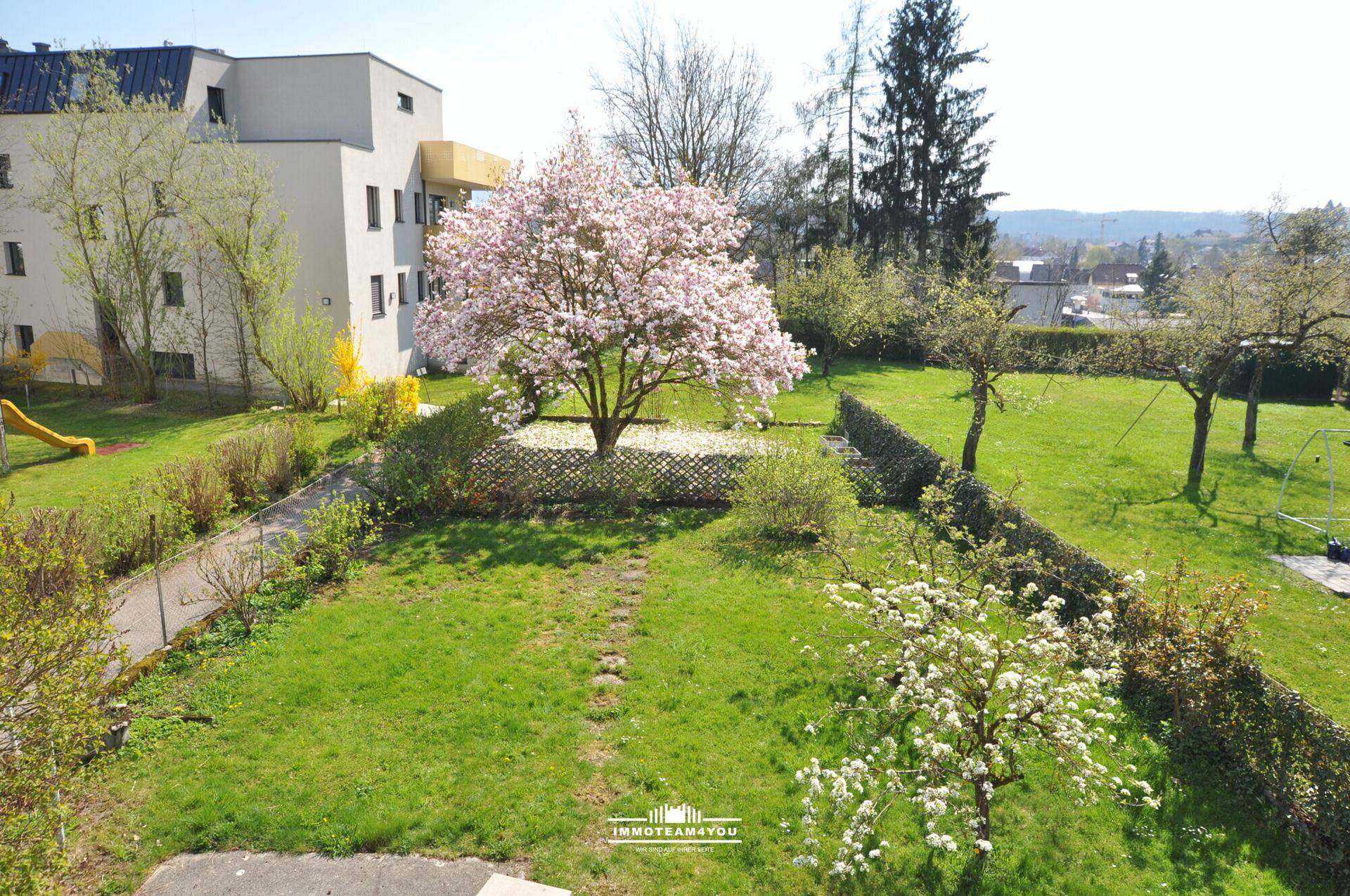 Großer Gemeinschaftsgarten mit blühendem Baum, Rasenfläche und Kinderspielgeräten.