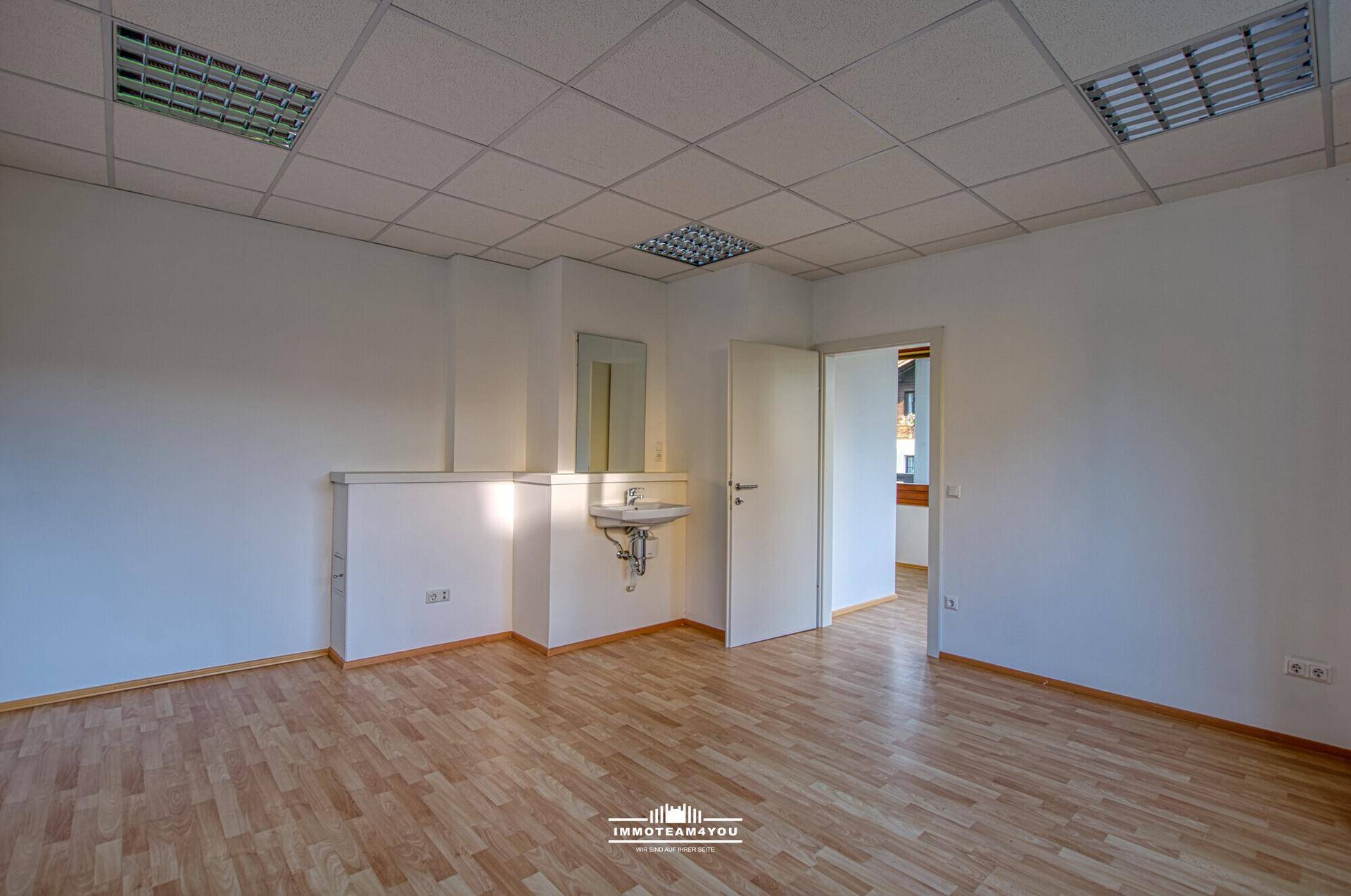 Geräumiger Büroraum mit Laminatboden, Deckenleuchten und einem kleinen Waschbeckenbereich an der Wand.
