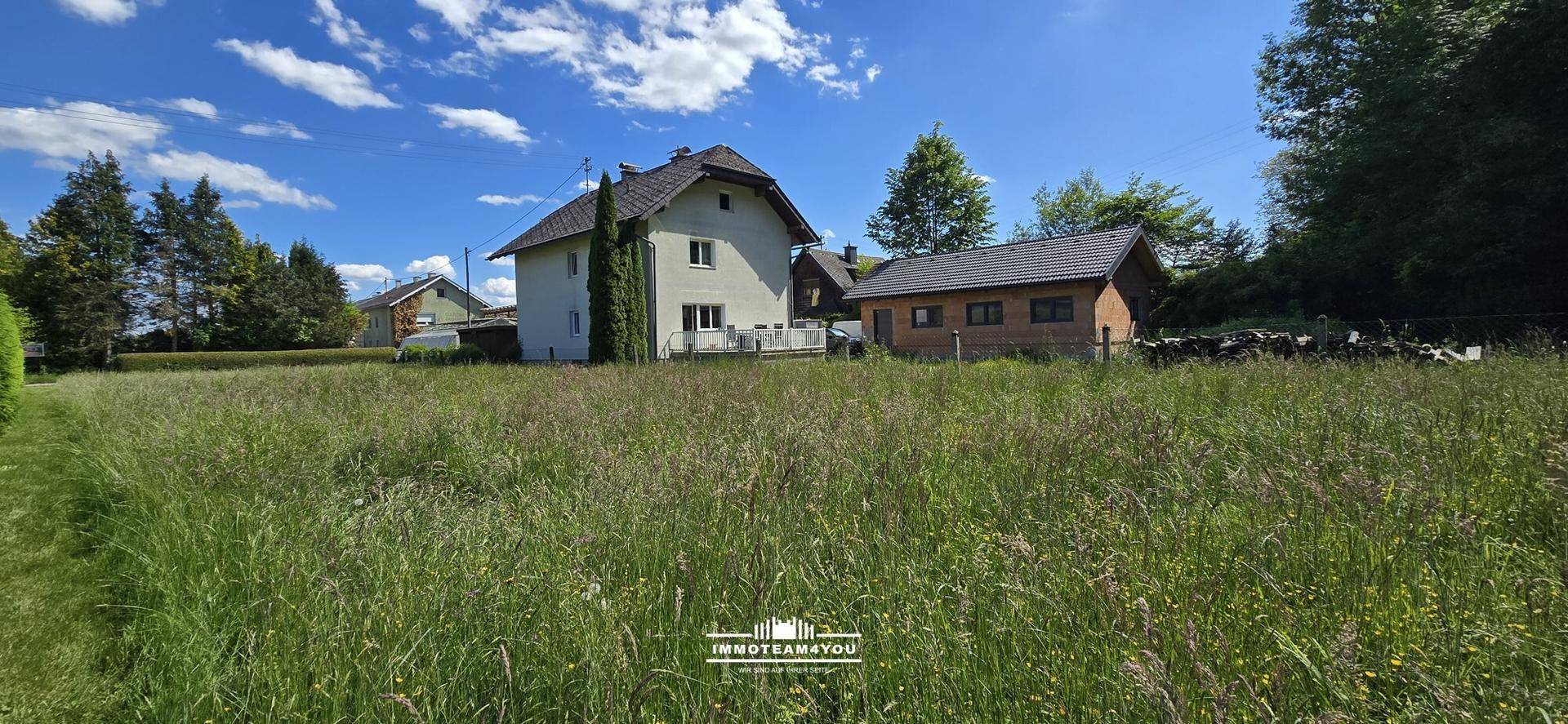 Weitläufige, überwachsene Rasenfläche mit einem Wohnhaus und einem Nebengebäude an einem sonnigen Tag.