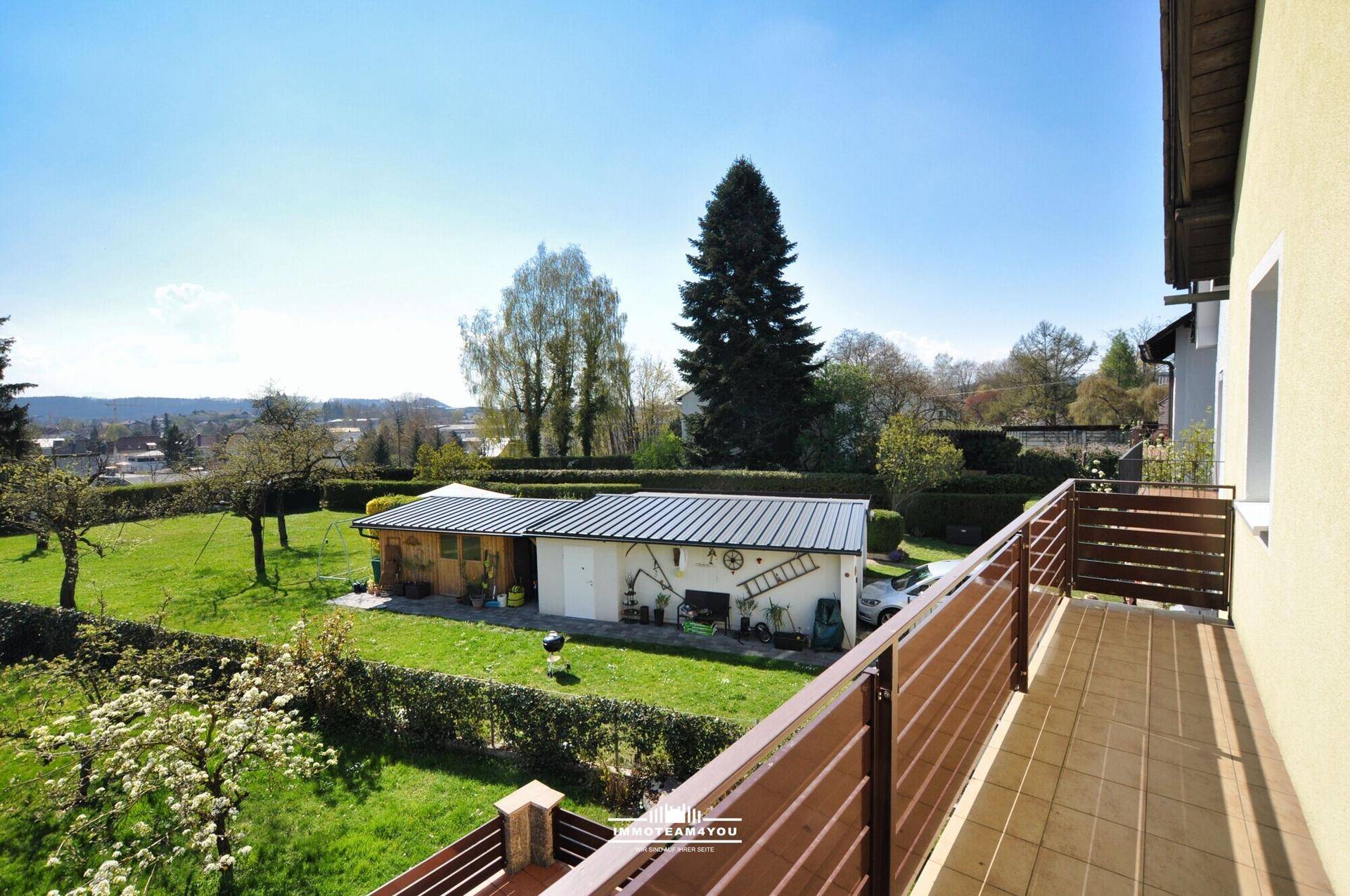 Balkon mit Weitblick über grüne Landschaft und Nebengebäude unter blauem Himmel.