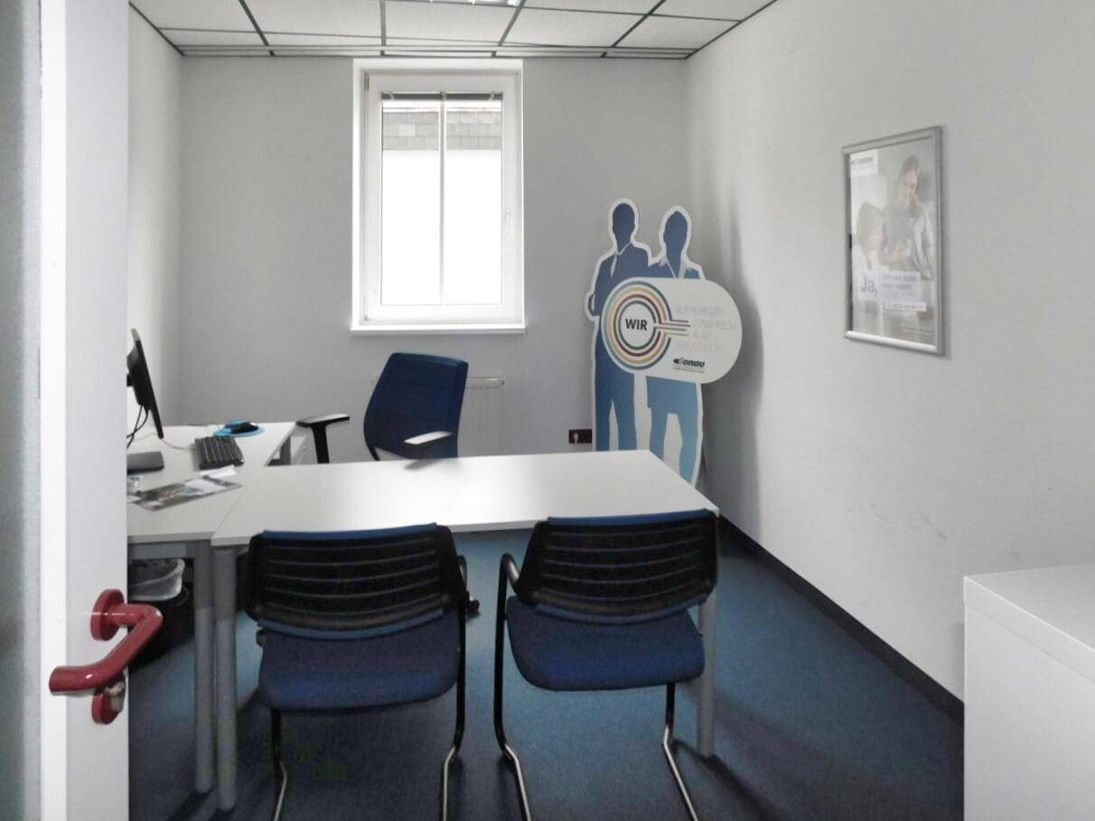 Helles Büro mit einem Fenster, Schreibtisch und blauen Stühlen, geeignet für Einzel- oder Teamarbeit.