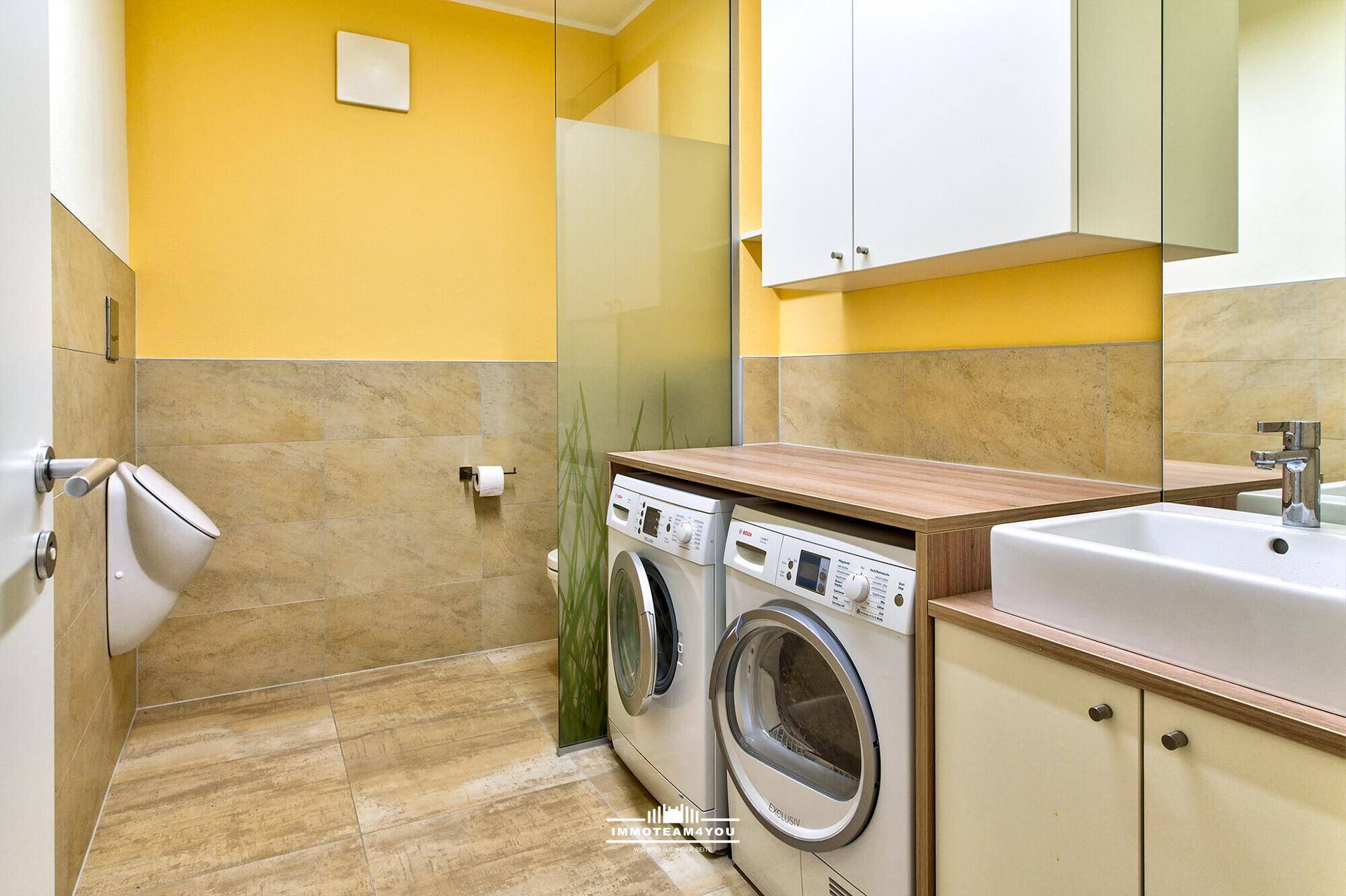 Funktionales Badezimmer mit Urinal, Waschmaschine, Trockner und Waschbecken in Gelb- und Beigetönen.