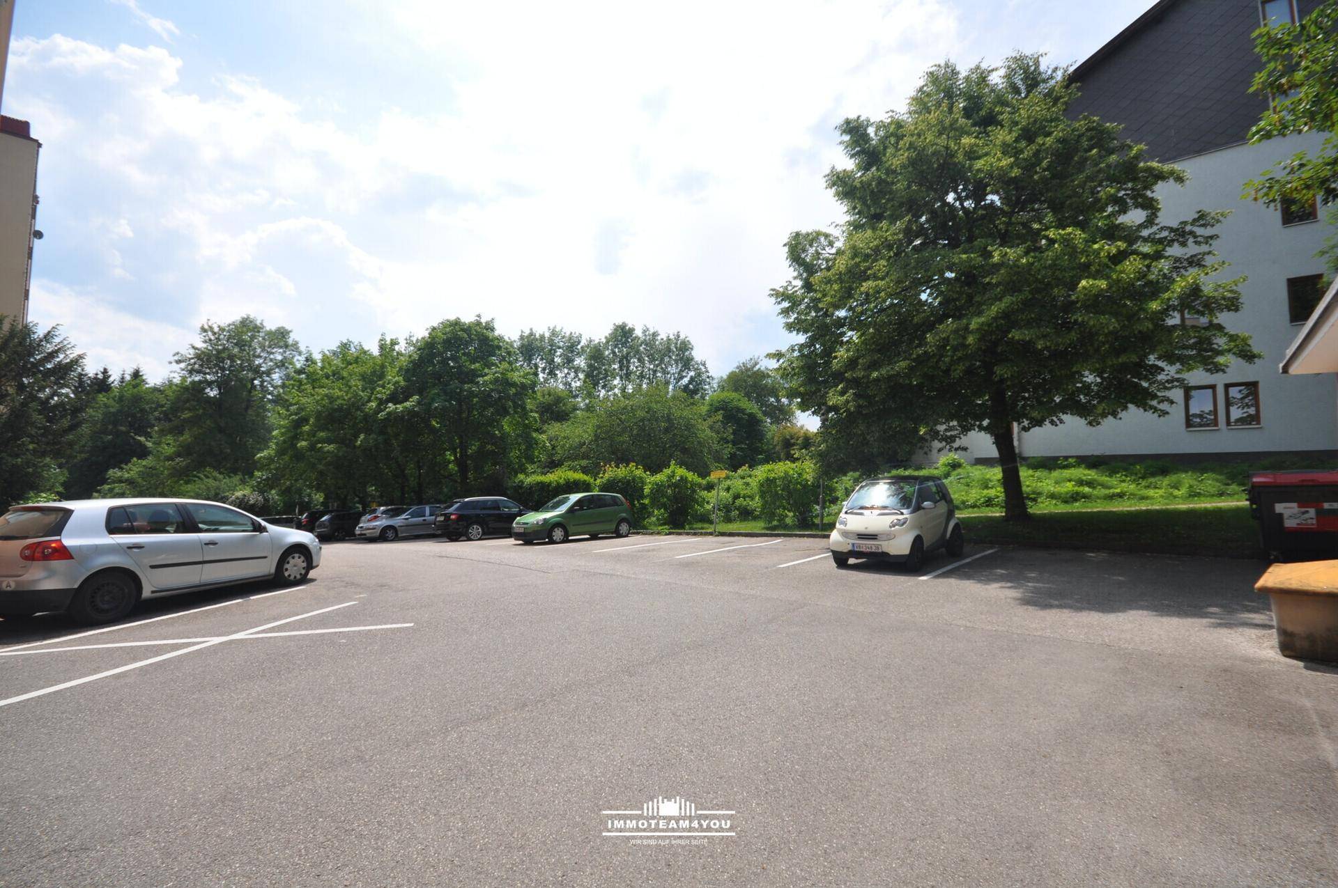 Außenparkplatz mit mehreren Fahrzeugen und umgebenden Bäumen an einem sonnigen Tag.