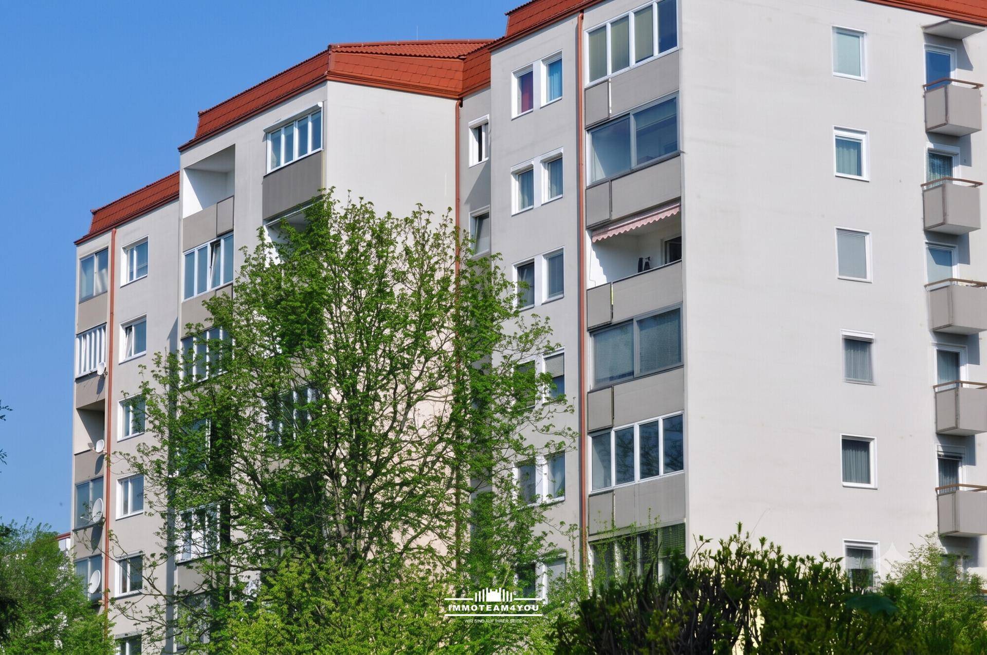 Fassade eines Mehrfamilienhauses mit Balkonen und großen Fenstern, umgeben von Bäumen.