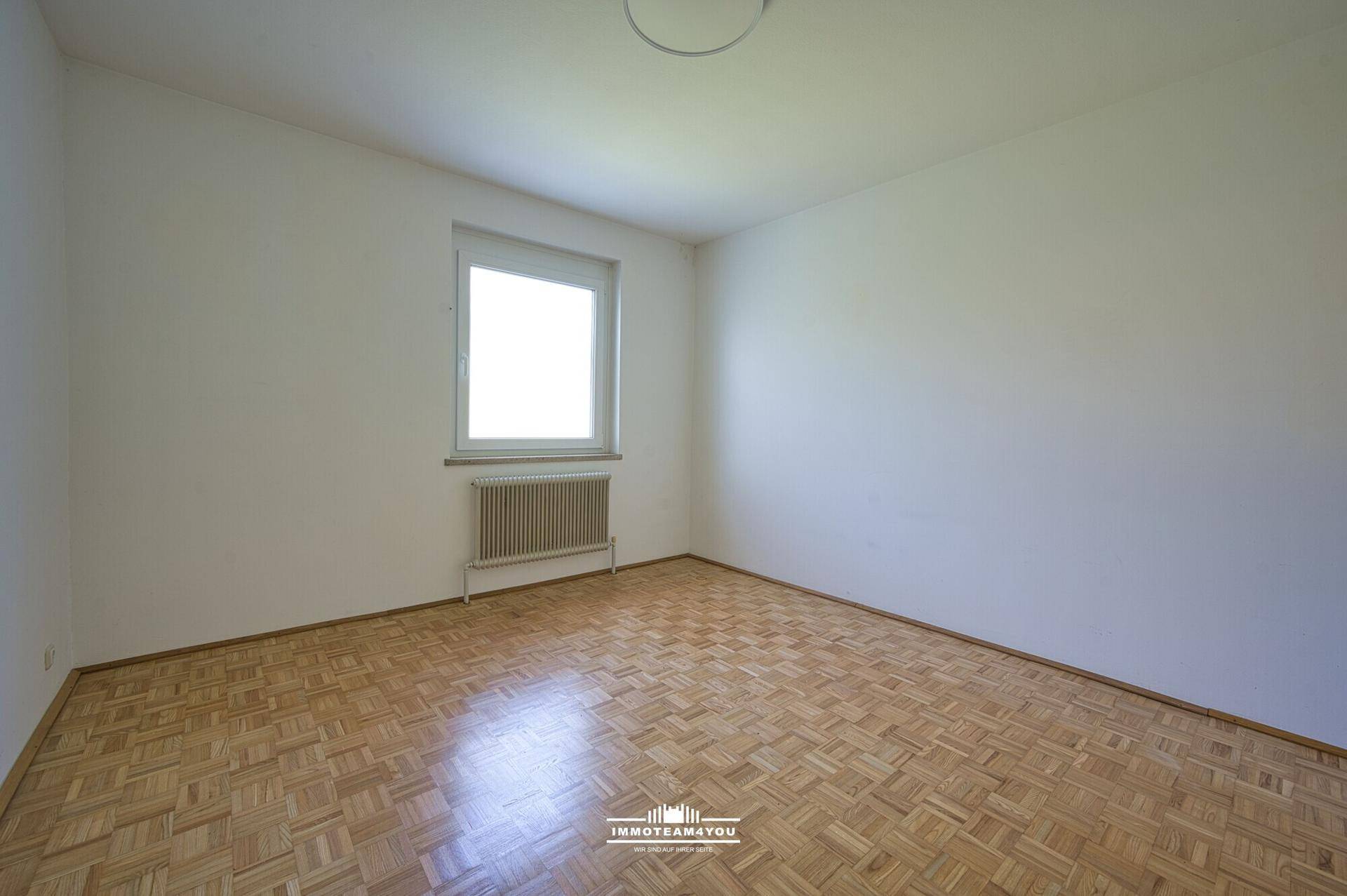 Leeres Schlafzimmer mit Parkettboden, großem Fenster und Heizkörper, bereit zur individuellen Gestaltung.