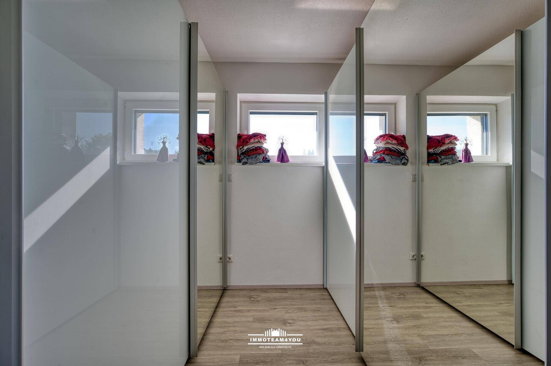 Begehbarer Kleiderschrank mit Spiegeltüren und Fenster, auf hellem Holzboden.