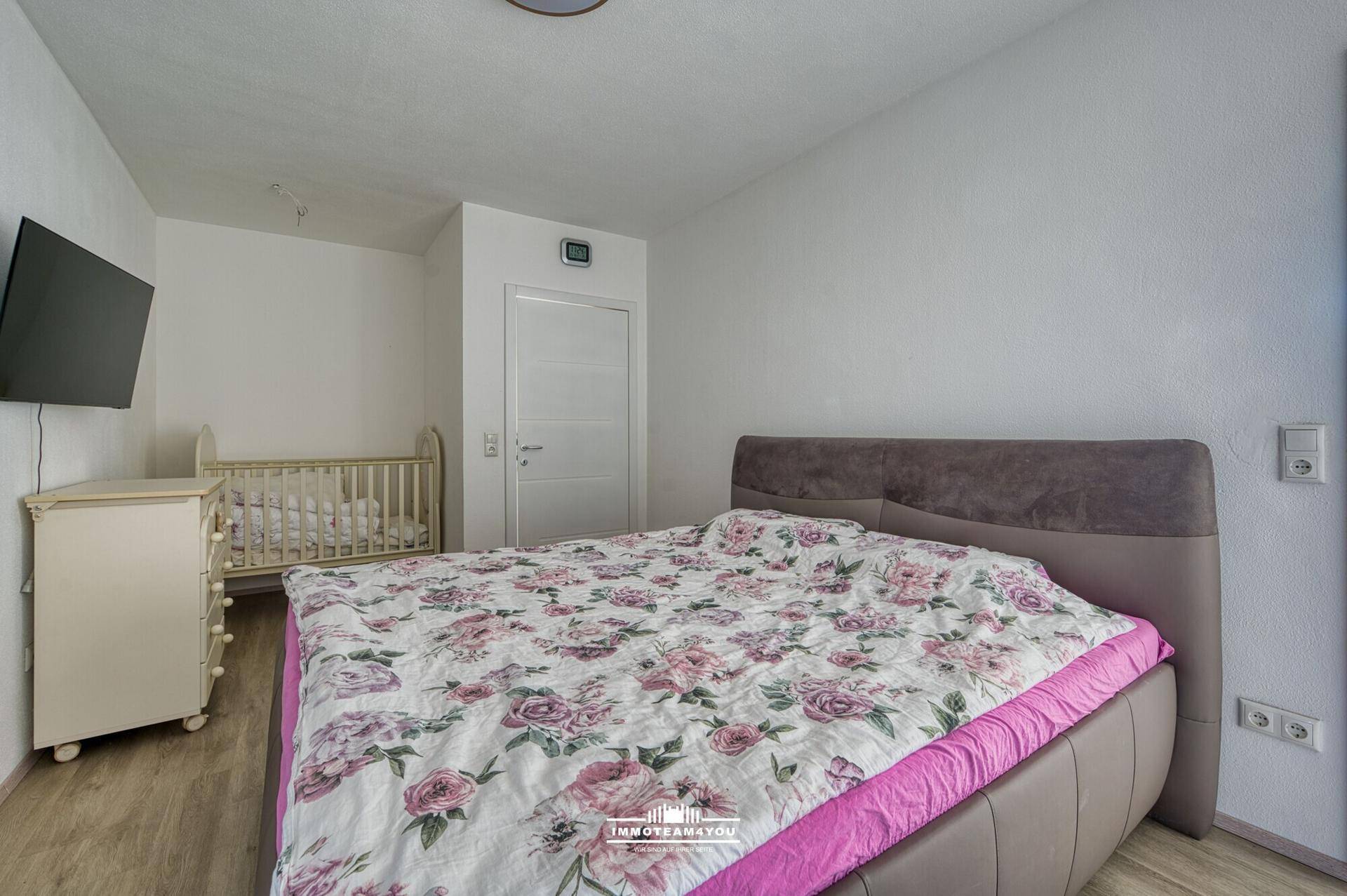 Schlafzimmer mit Doppelbett, floraler Bettwäsche und einem Babybett, auf hellem Holzboden.