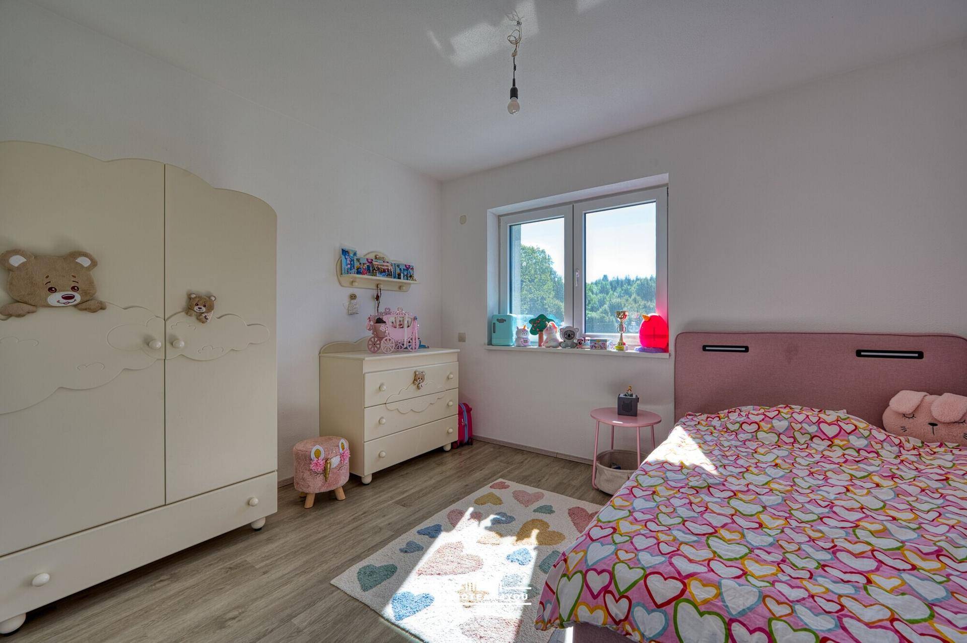 Kinderzimmer mit rosa Bett, weißem Kleiderschrank und Blick ins Grüne, auf hellem Holzboden.