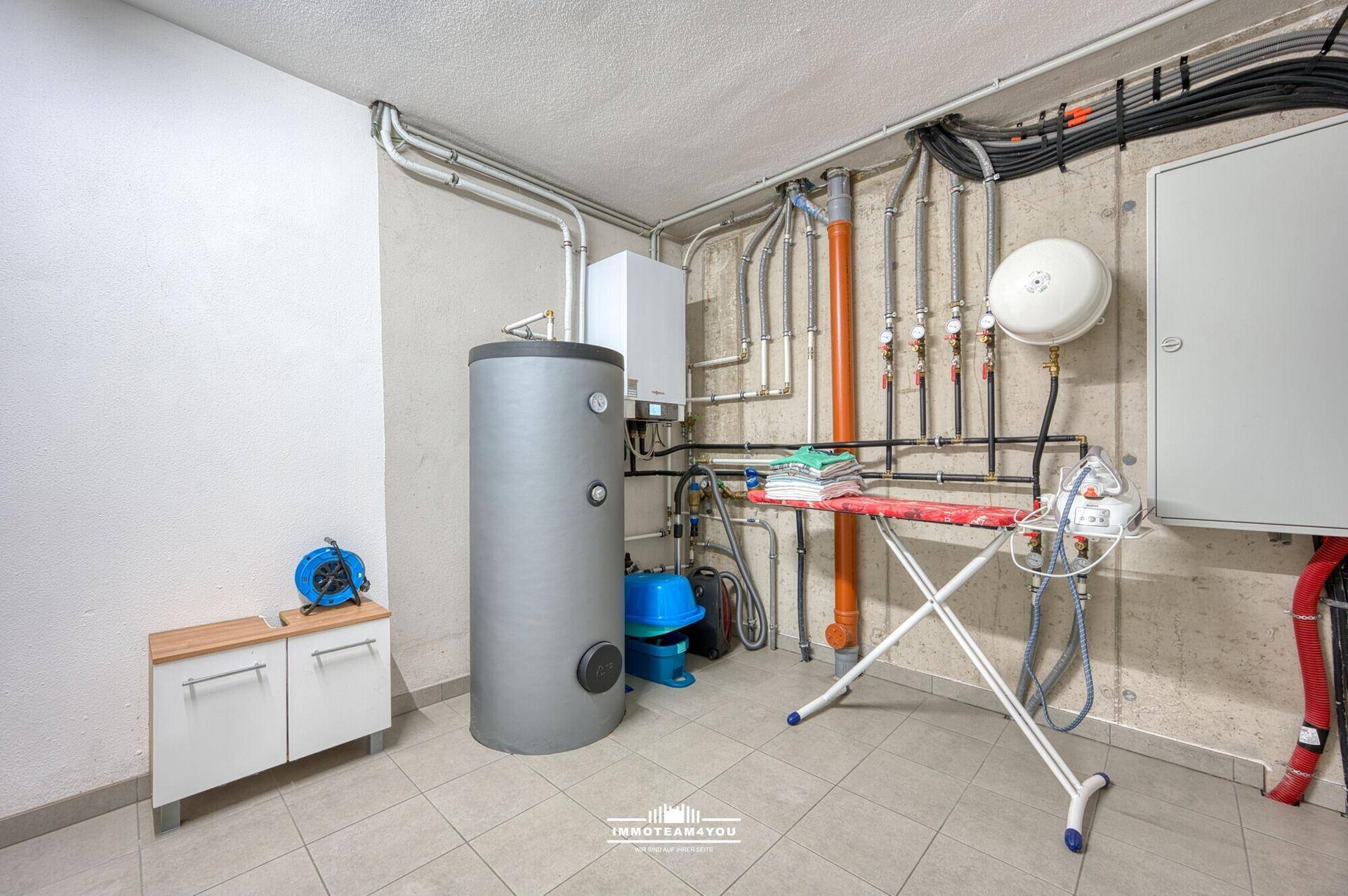 Technikraum mit Warmwasserspeicher, Leitungen und Bügelbrett, auf Fliesenboden.