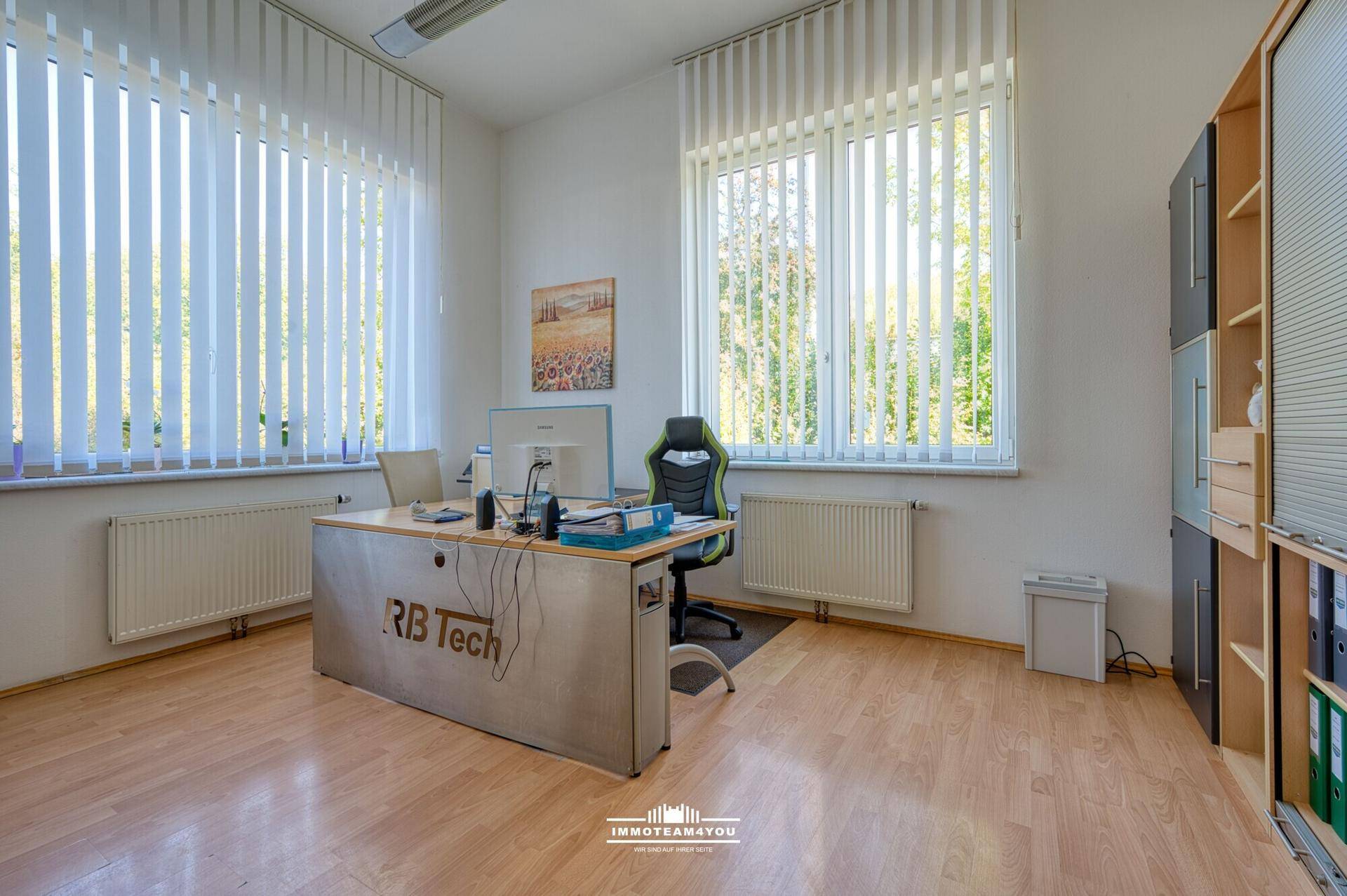 Helles Büro mit Holzfußboden, großen Fenstern und vertikalen Jalousien.