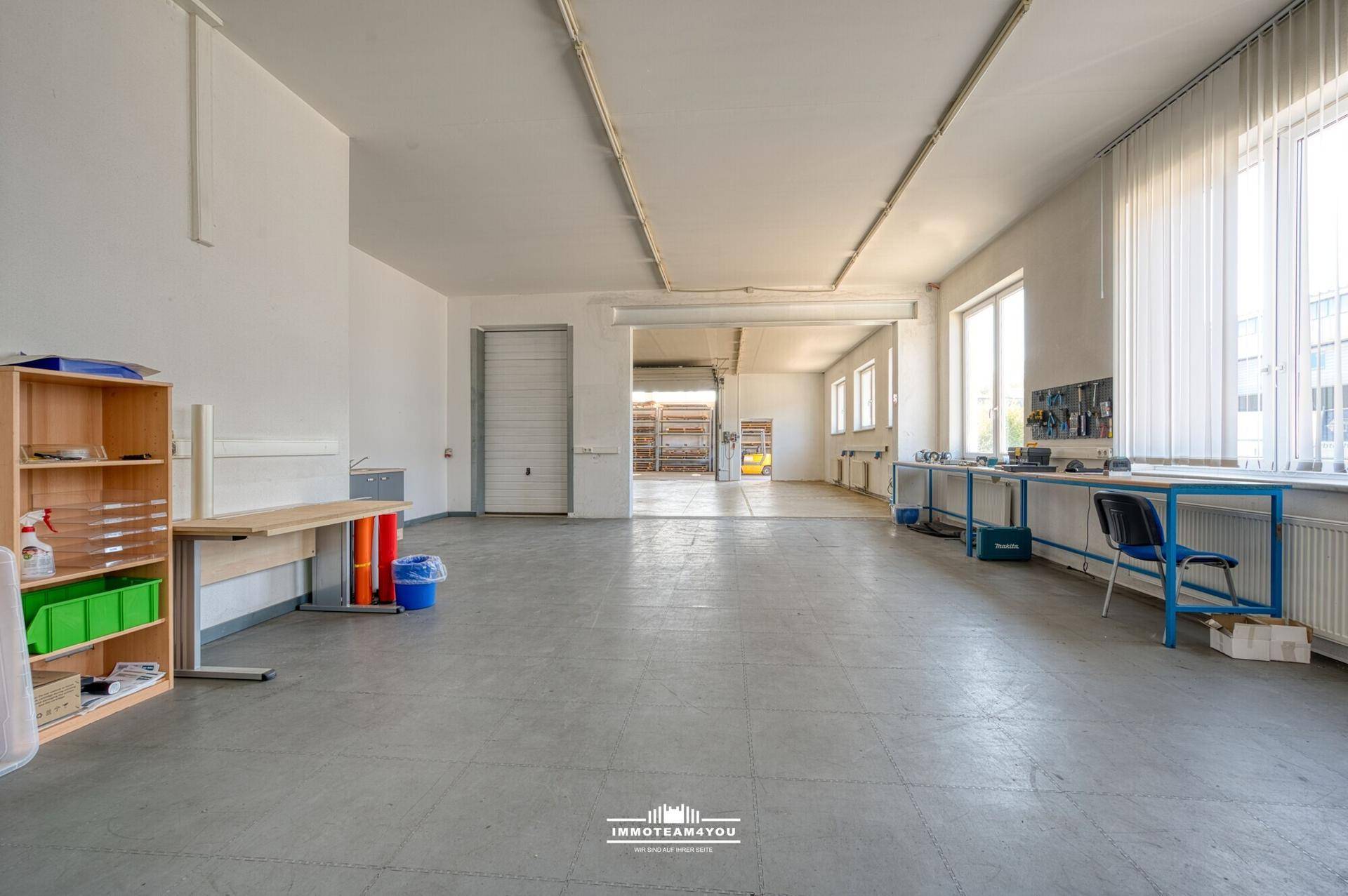 Geräumiger Lagerbereich mit Betonboden, Rolltor und viel Tageslicht durch die Fenster.