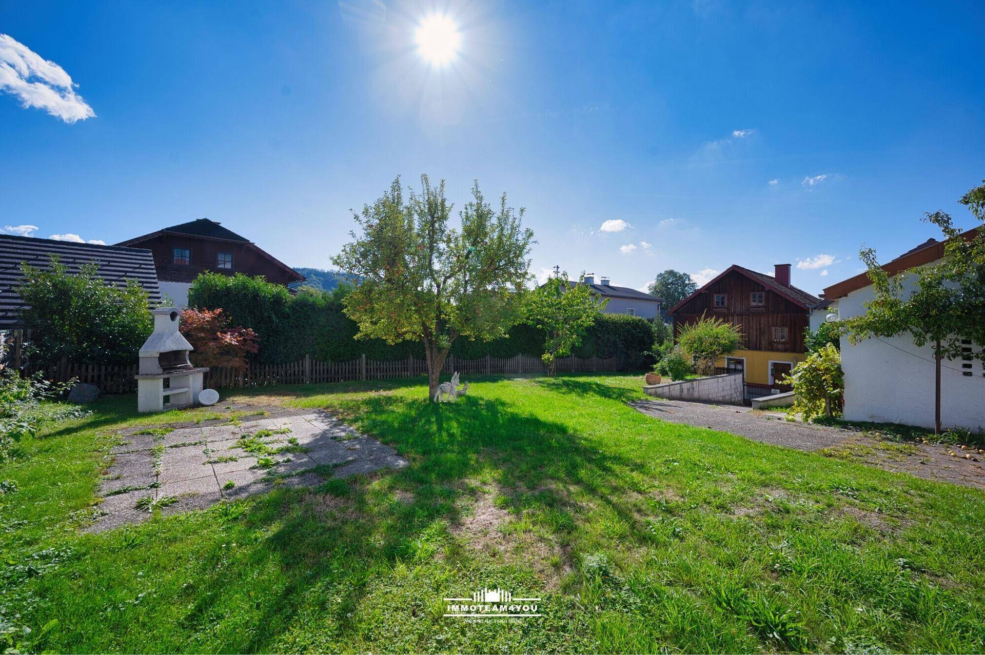 Sonniger Garten mit Rasen, Obstbäumen und gemauertem Grill.