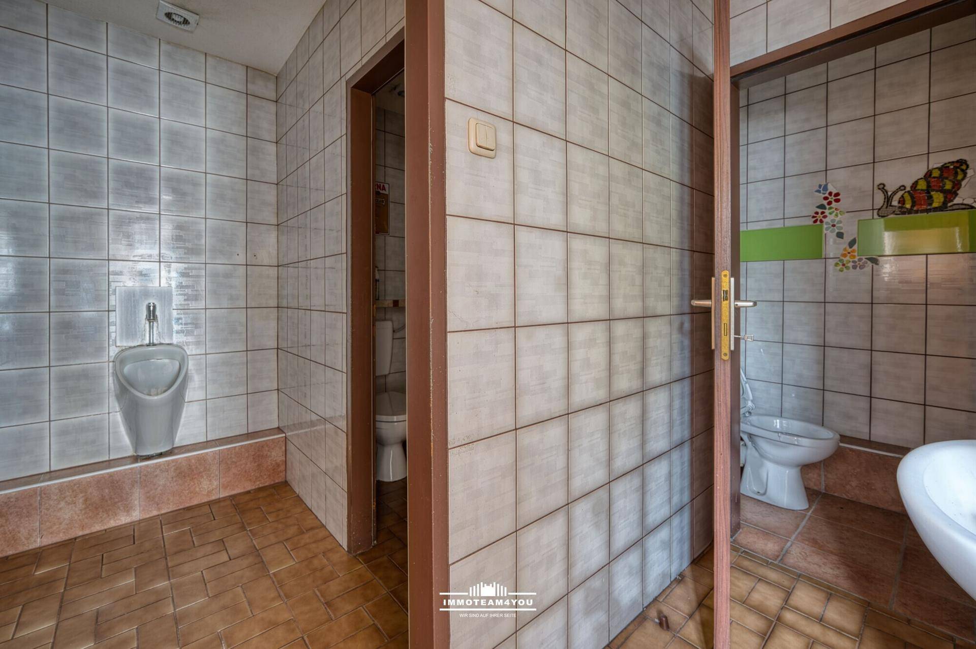 Mehrere Toilettenräume mit Fliesenboden und Urinalen, in älterem, aber funktionsfähigem Zustand.
