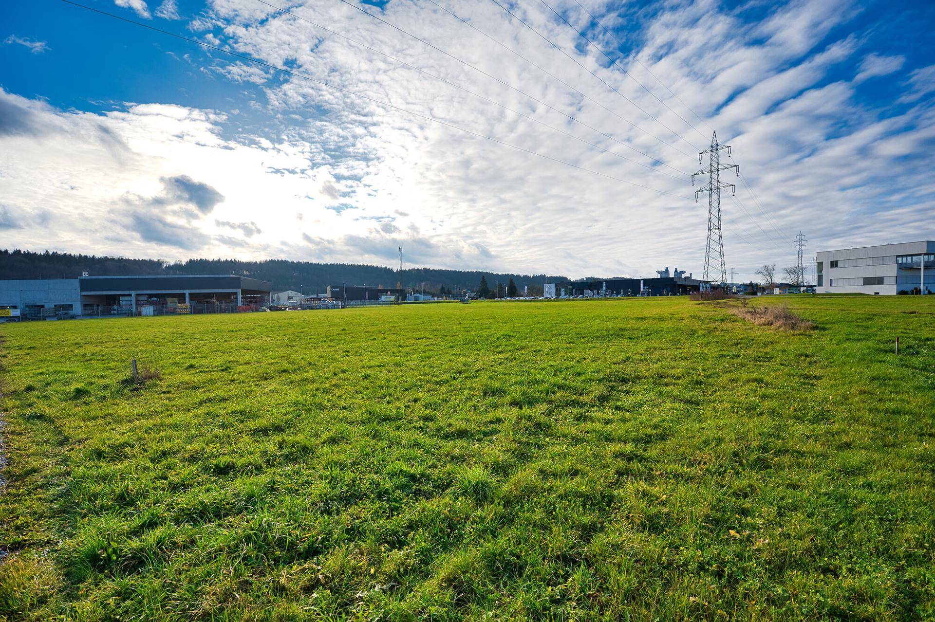 Ausgedehntes grünes Feld mit Blick auf Industriegebäude und einen hohen Strommast unter einem teilweise bewölkten Himmel.
