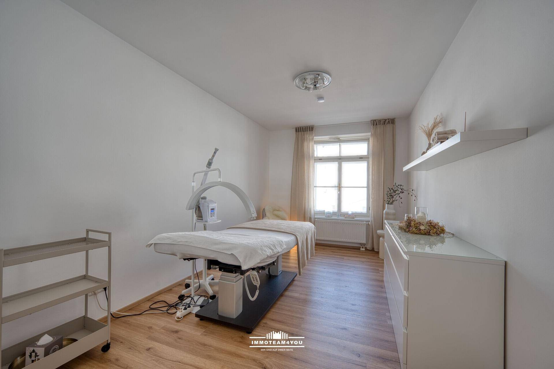 Behandlungsraum mit Liege, Fenster und Holzfußboden, hell und funktional eingerichtet.