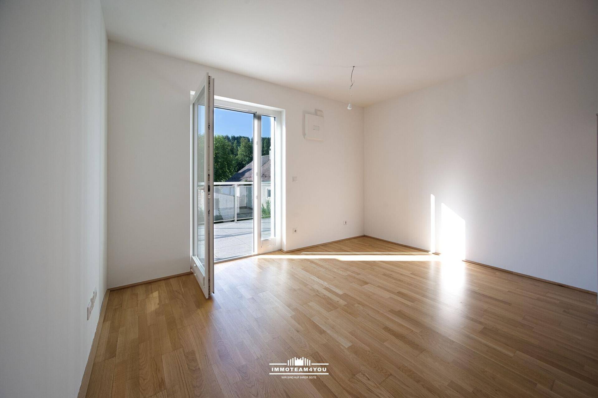 Lichtdurchfluteter Wohnraum mit hellem Parkettboden und offener Balkontür, die eine grüne Aussicht freigibt.
