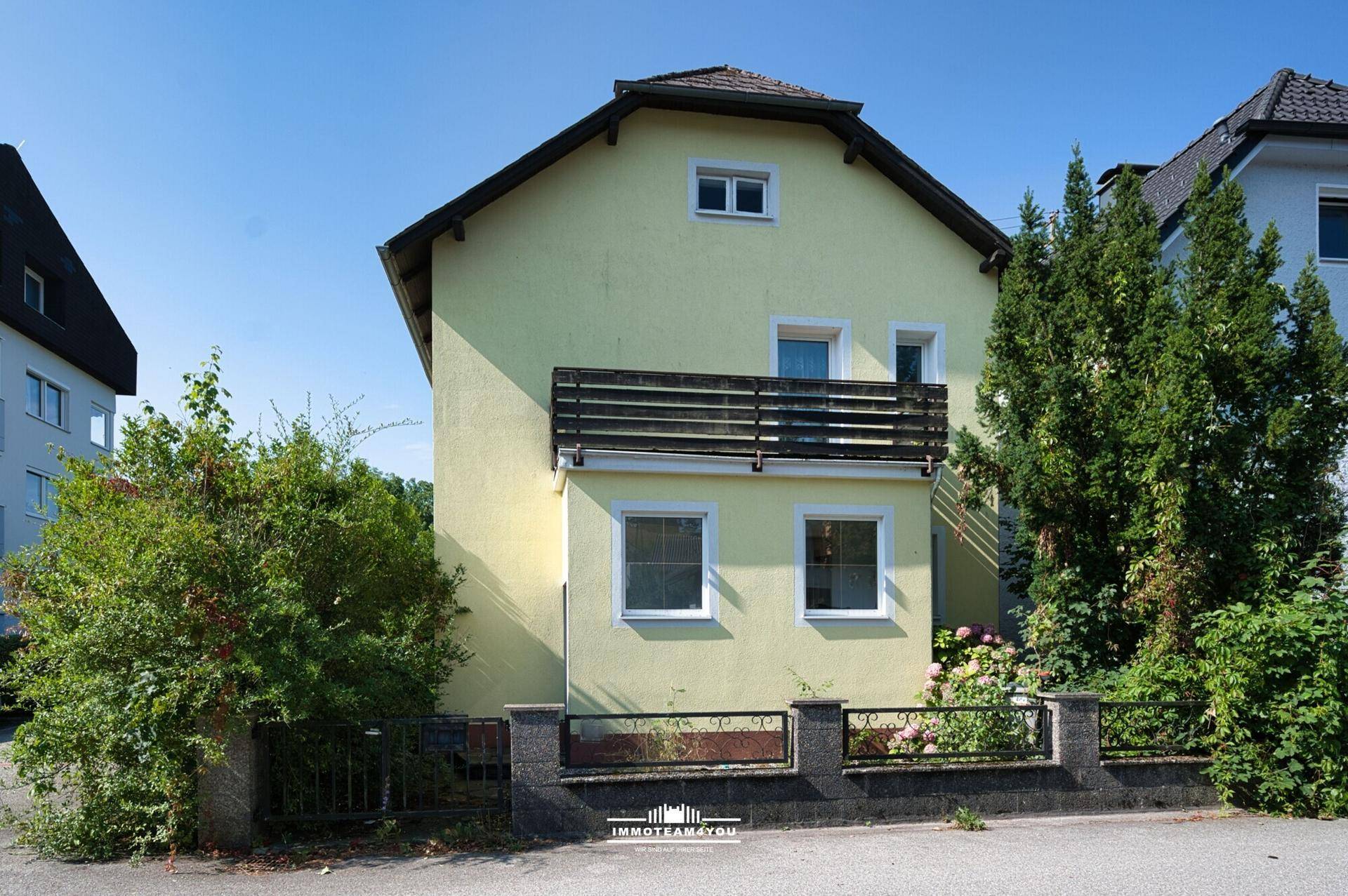 Rückansicht des gelben Hauses mit Balkon und üppiger Bepflanzung im Gartenbereich.