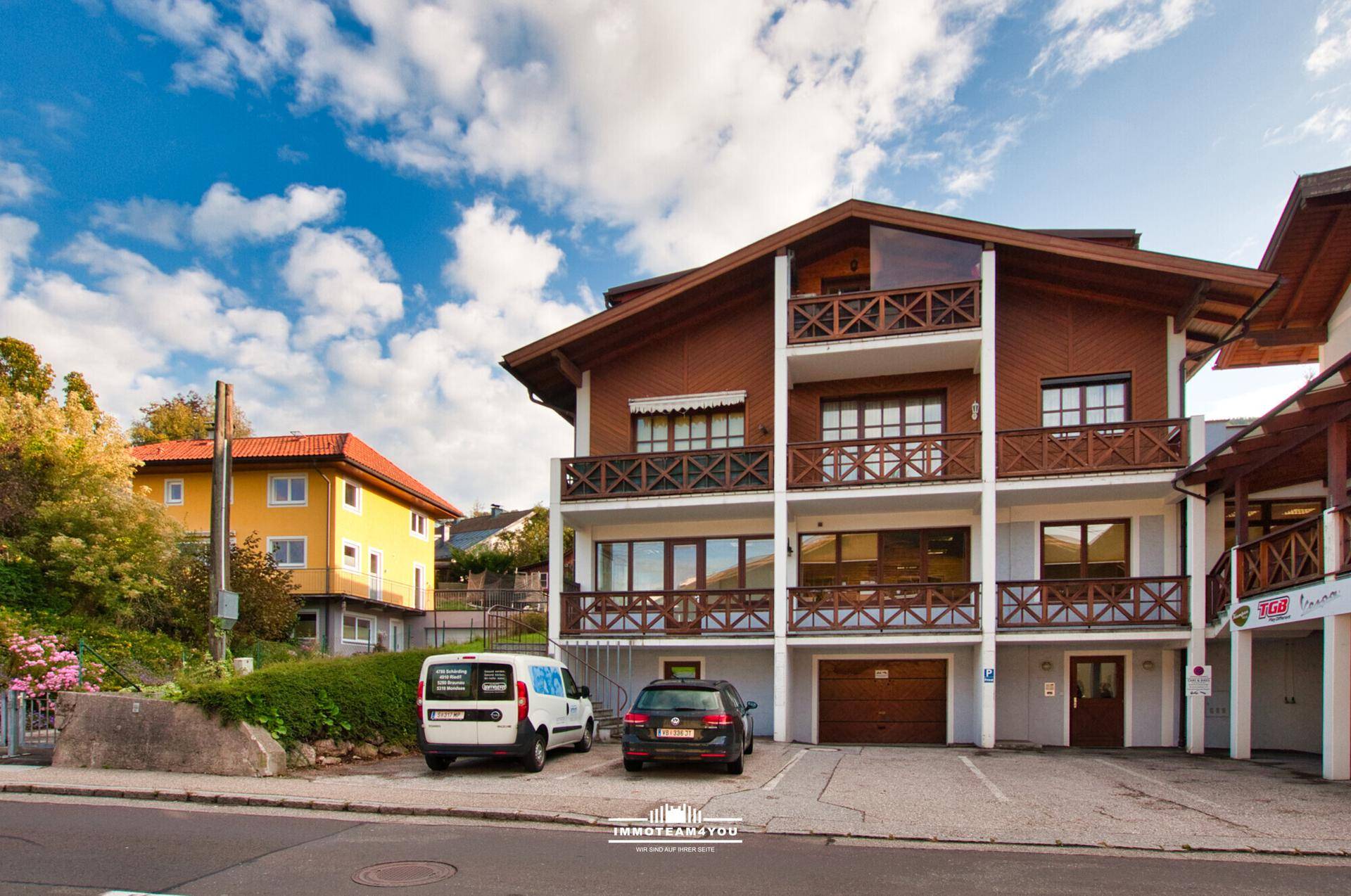 Mehrstöckiges Gebäude mit Holzfassade, Balkonen und Parkplätzen vor dem Eingang unter blauem Himmel.