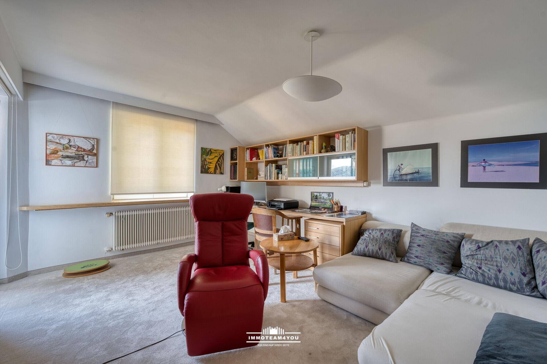 Helles Wohnzimmer mit Teppichboden, Bücherregal und bequemen Sitzgelegenheiten.