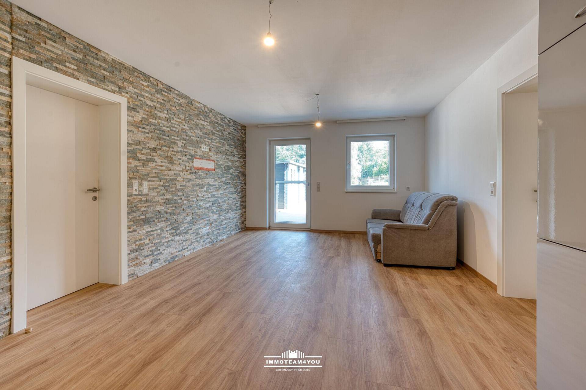 Leeres Wohnzimmer mit Laminatboden, Steinwand und Zugang zu einem Außenbereich.